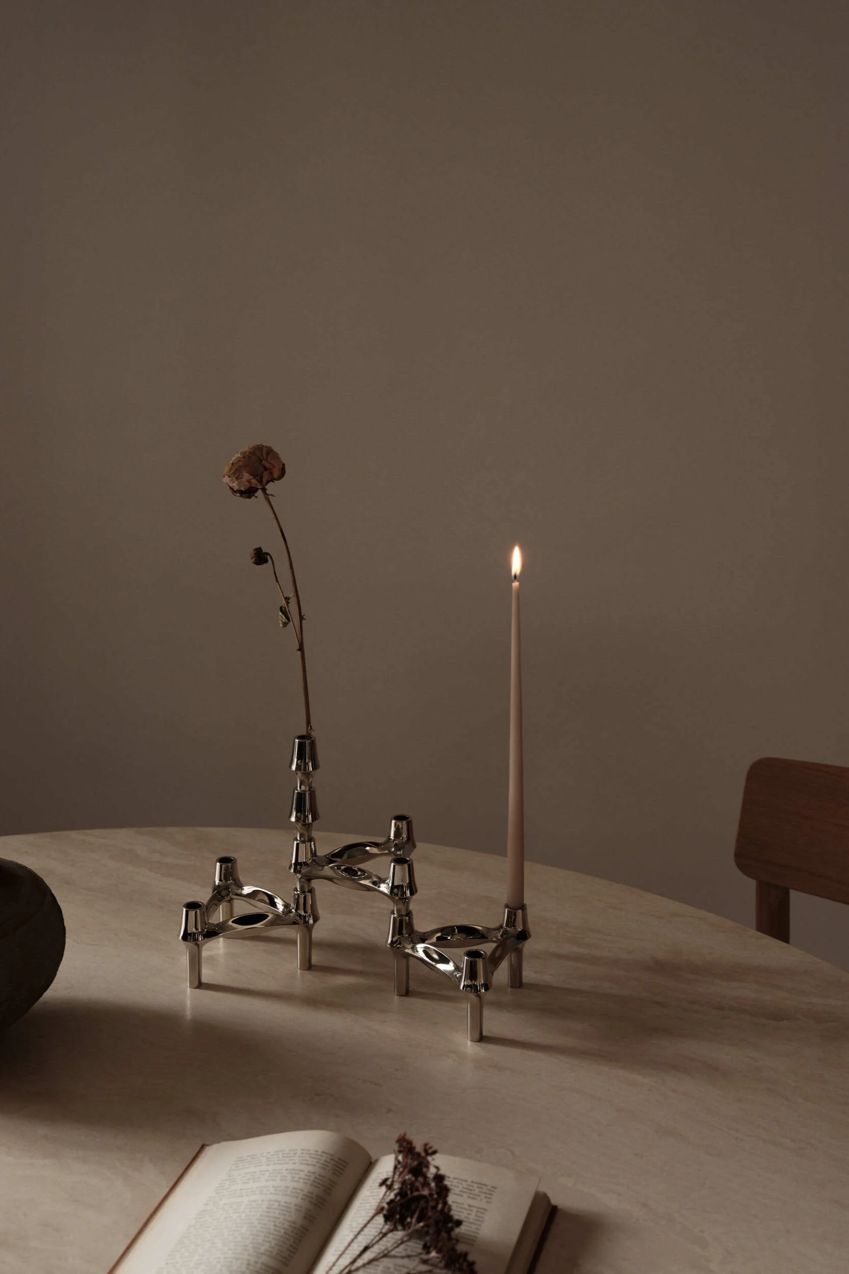 Deux bougeoirs modulaires argentés posés sur une table ronde, l'un avec une bougie allumée, l'autre avec une fleur séchée. Un livre ouvert est visible au premier plan.