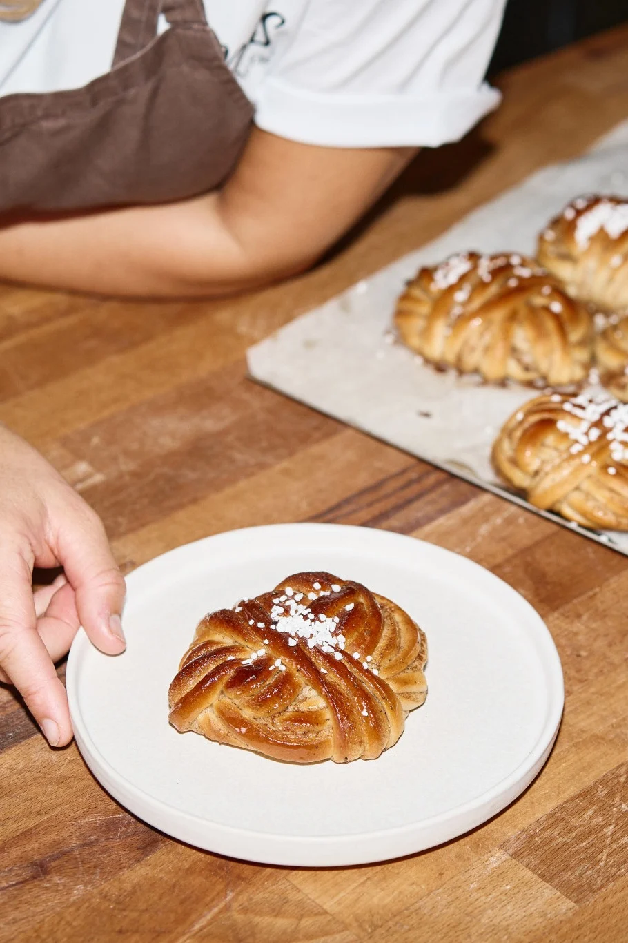Une main pose une brioche à la cannelle tressée et saupoudrée de sucre sur une assiette; d'autres brioches sur un plateau.