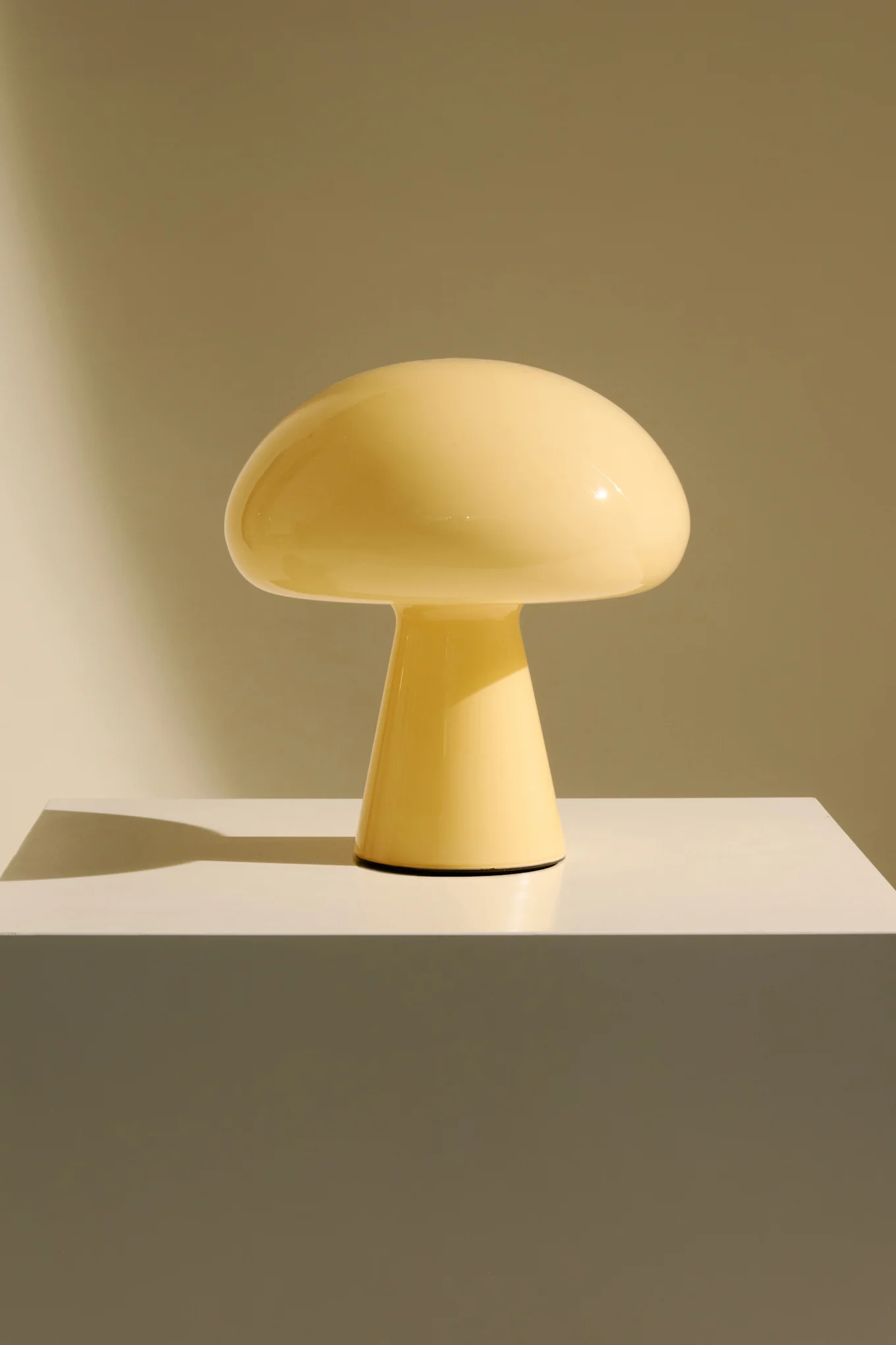 Une lampe jaune pâle en forme de champignon sur un piédestal blanc, fond brun clair.