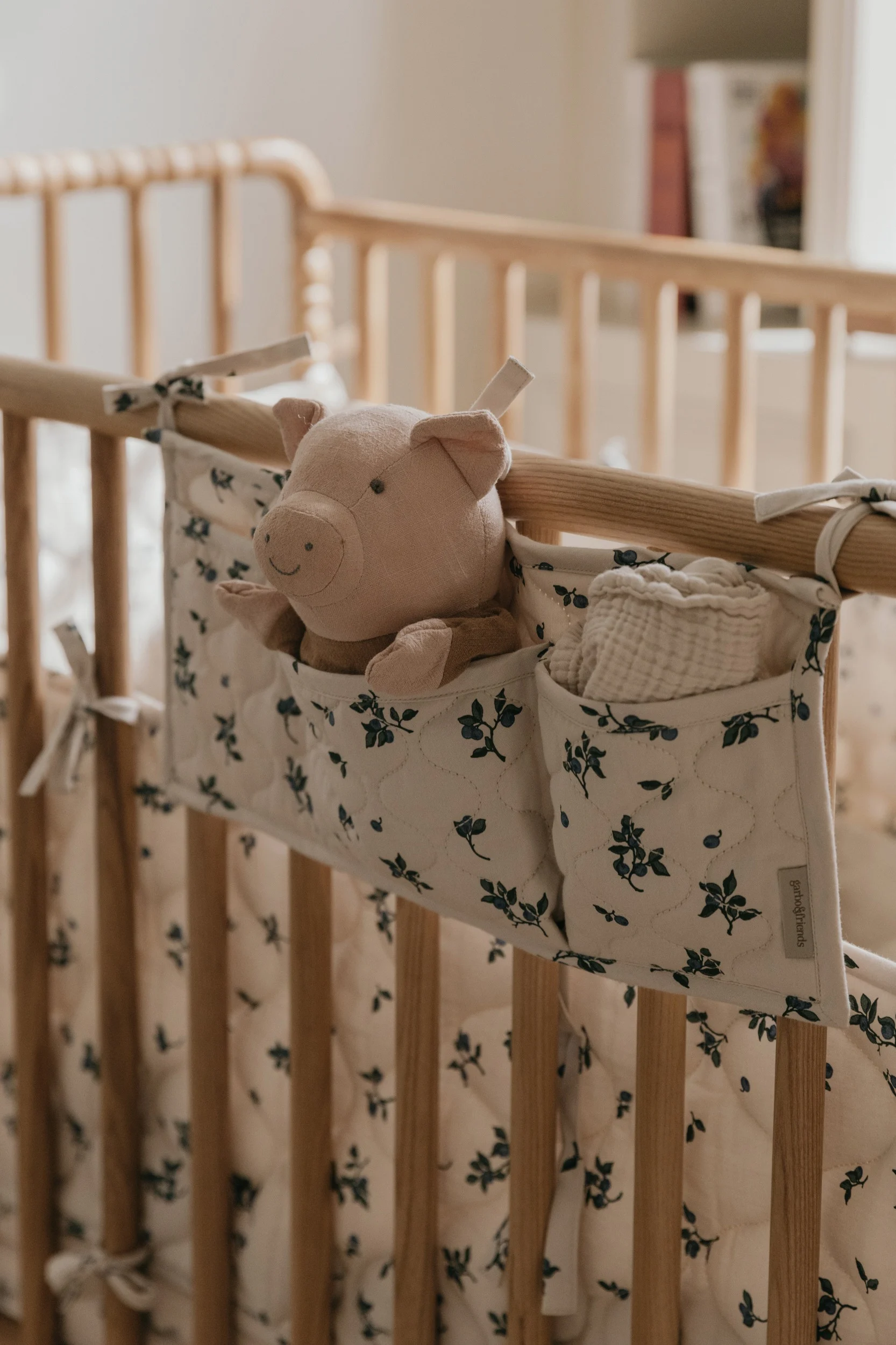 Un cochonnet en peluche rose et un tissu en mousseline blanc dans un organisateur floral crème et bleu accroché à un lit bébé en bois.