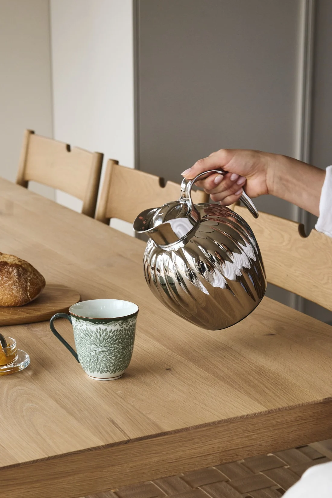 Une main verse d'une carafe thermique argentée cannelée dans une tasse verte sur une table en bois avec du pain et confiture.