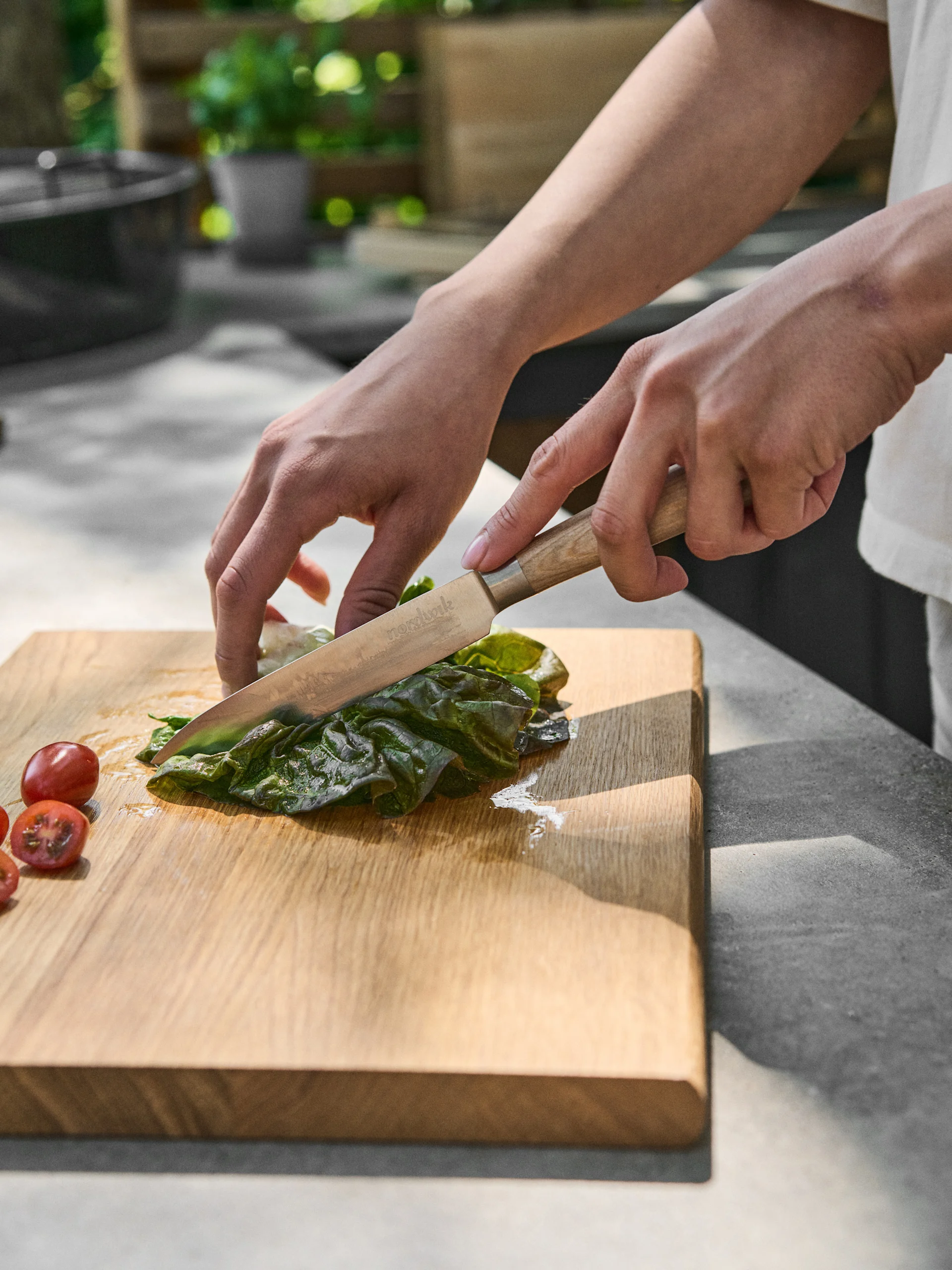 Mains coupant une feuille verte avec un couteau sur une planche en bois, à côté de tomates cerises.