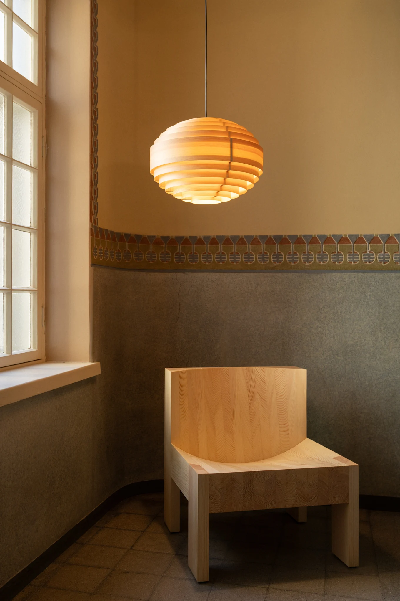 Suspension en lattes de bois au-dessus d'une chaise en bois clair dans une pièce aux murs décorés et fenêtre.