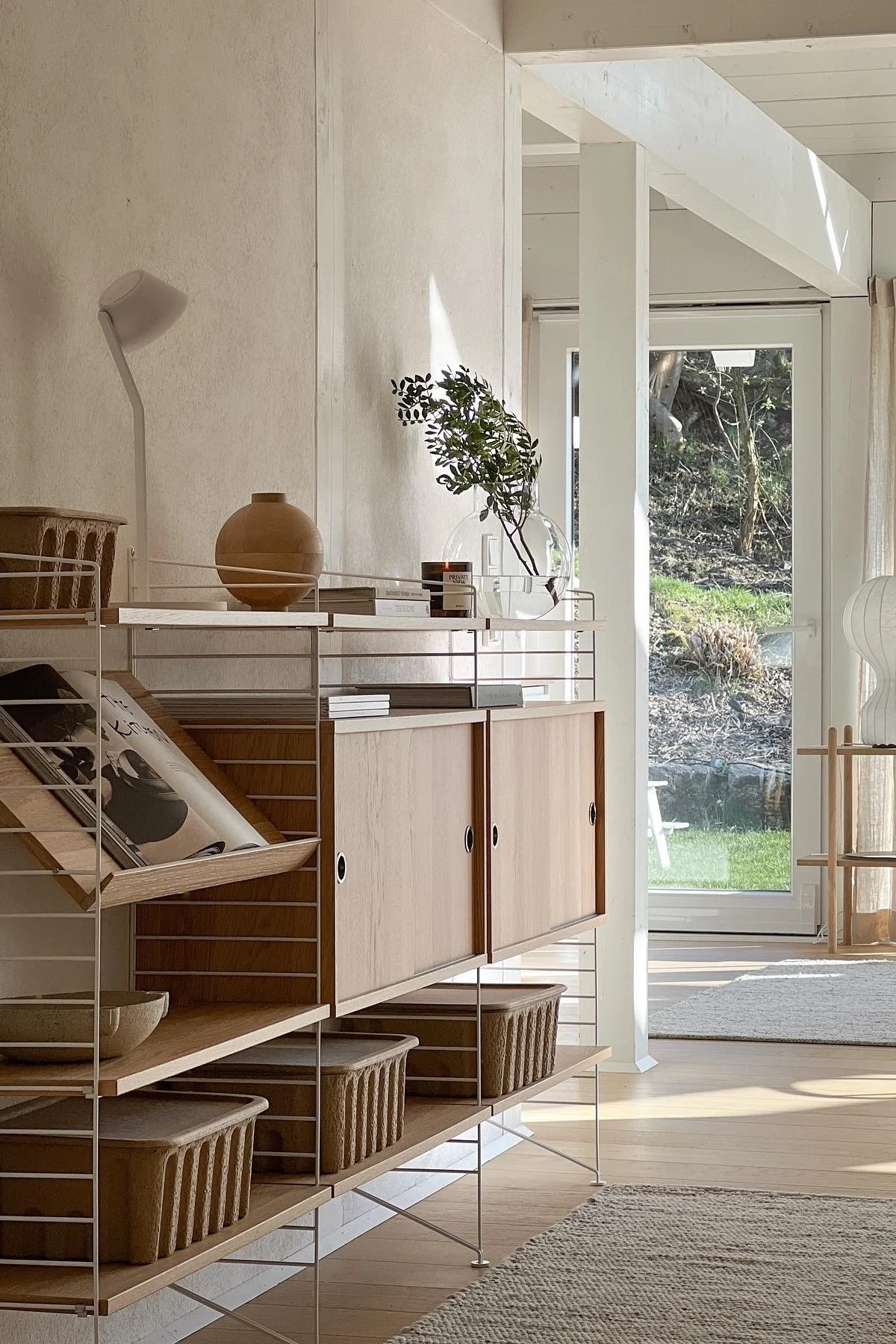 Pièce moderne et lumineuse avec étagère en bois clair et cadres blancs, contenant vase, livres, plantes et paniers, vue sur jardin.