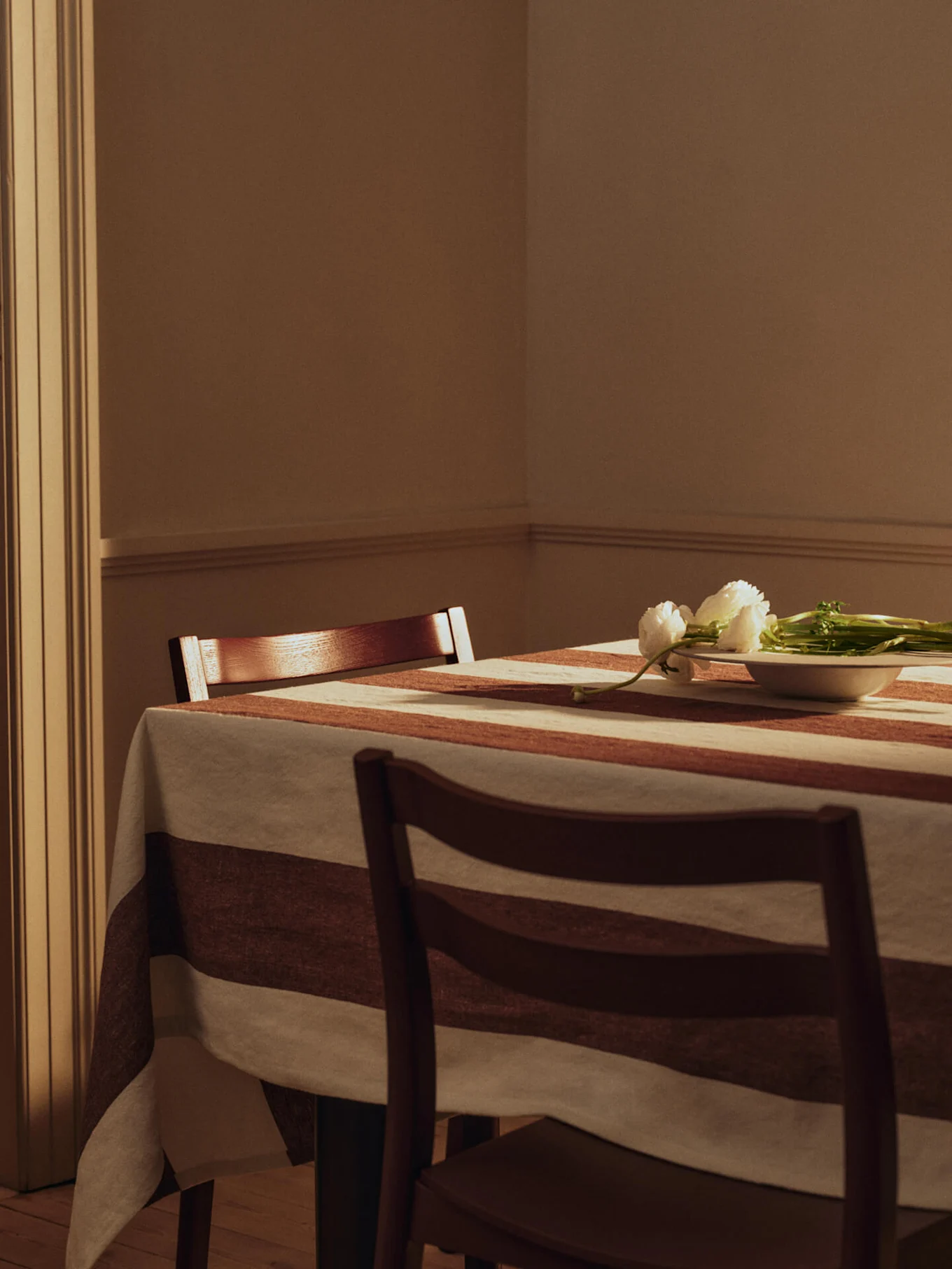 Table à manger avec nappe rayée, deux chaises en bois et fleurs blanches dans un petit bol.