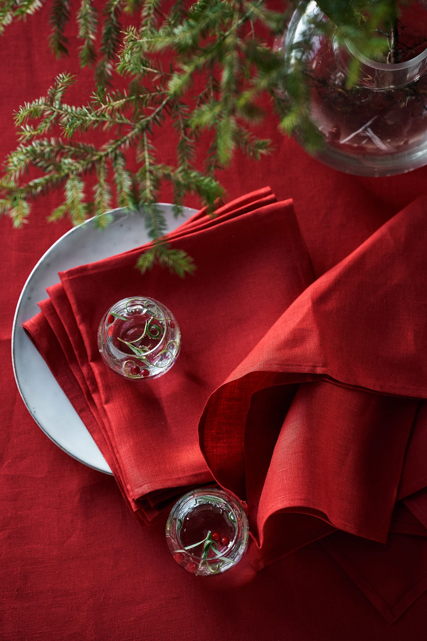 Table festive rouge avec des serviettes de table en lin rouges empilées sur une assiette, de petits bols en verre avec des canneberges et des branches de pin.