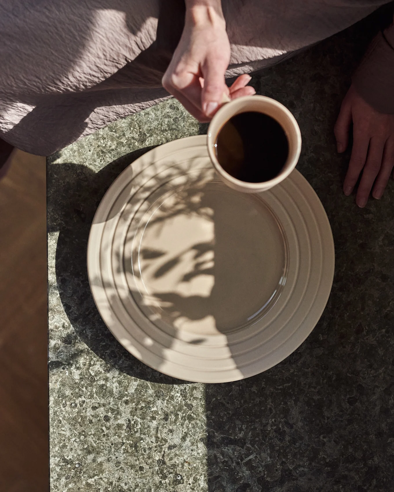 Vue plongeante : mains, l'une tient une tasse beige de café noir au-dessus d'une assiette beige sur une table en pierre foncée, avec des ombres de feuilles.