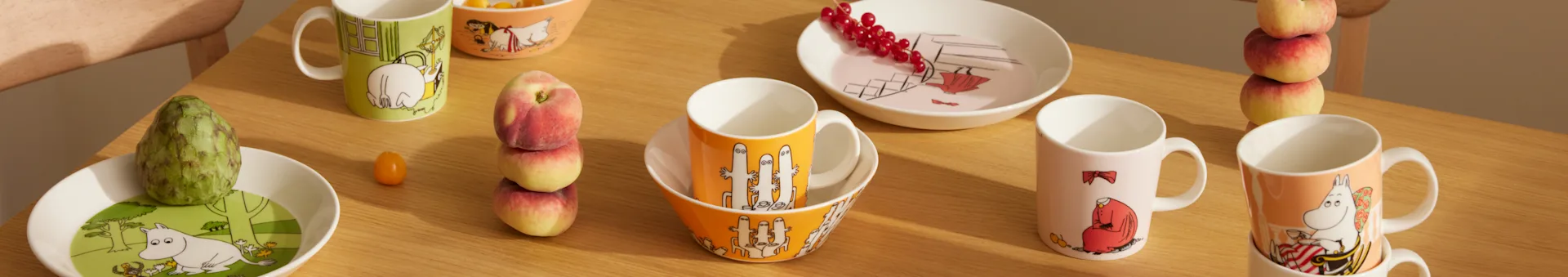 Vaisselle Moomin de couleurs et designs variés, tasses, bols et assiettes, avec des fruits sur une table en bois.