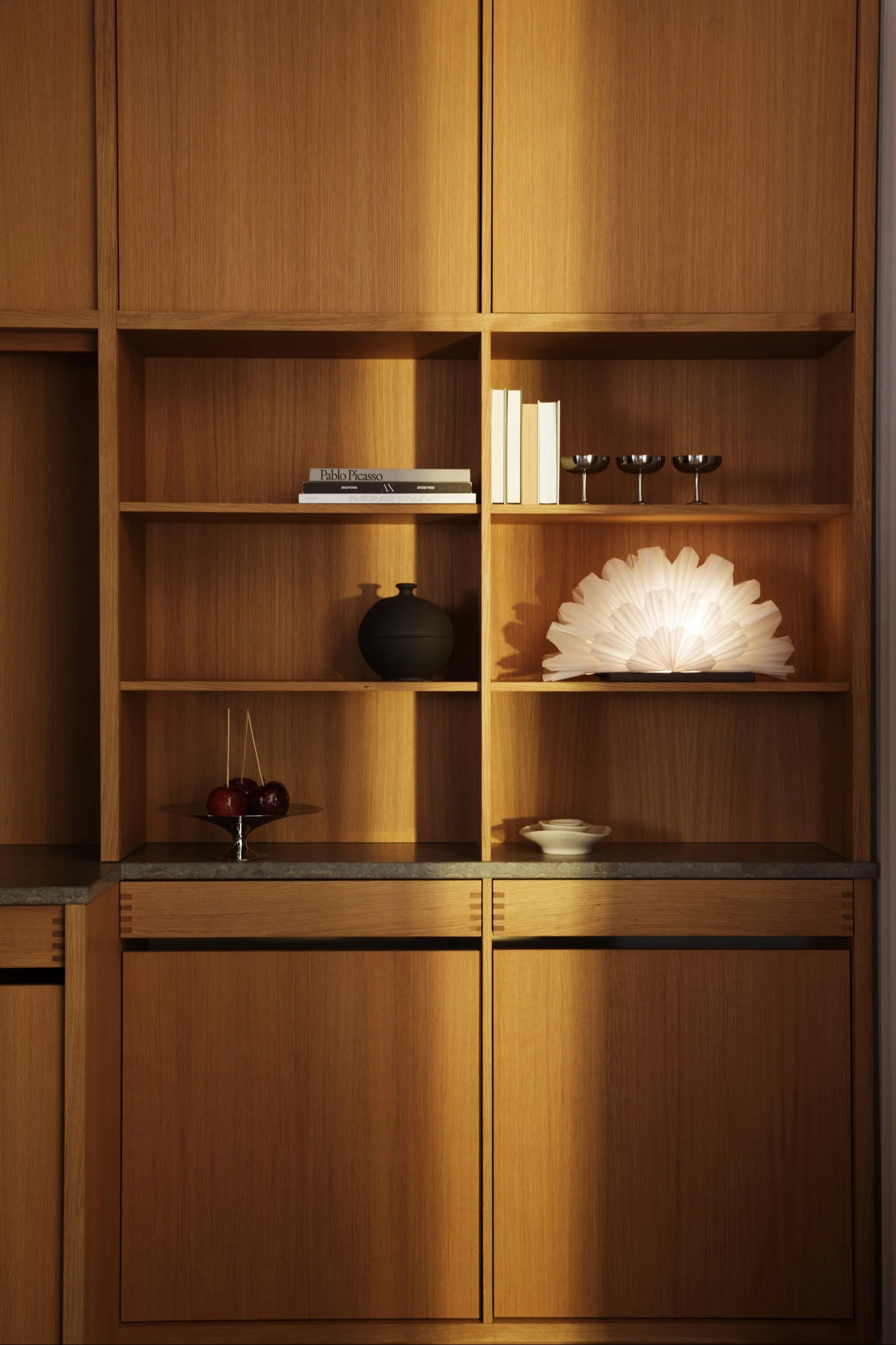 Un meuble en bois encastré moderne avec des étagères ouvertes présentant des livres, un vase noir, une lampe lumineuse en forme d'éventail, des pommes d'amour et des coupes argentées.