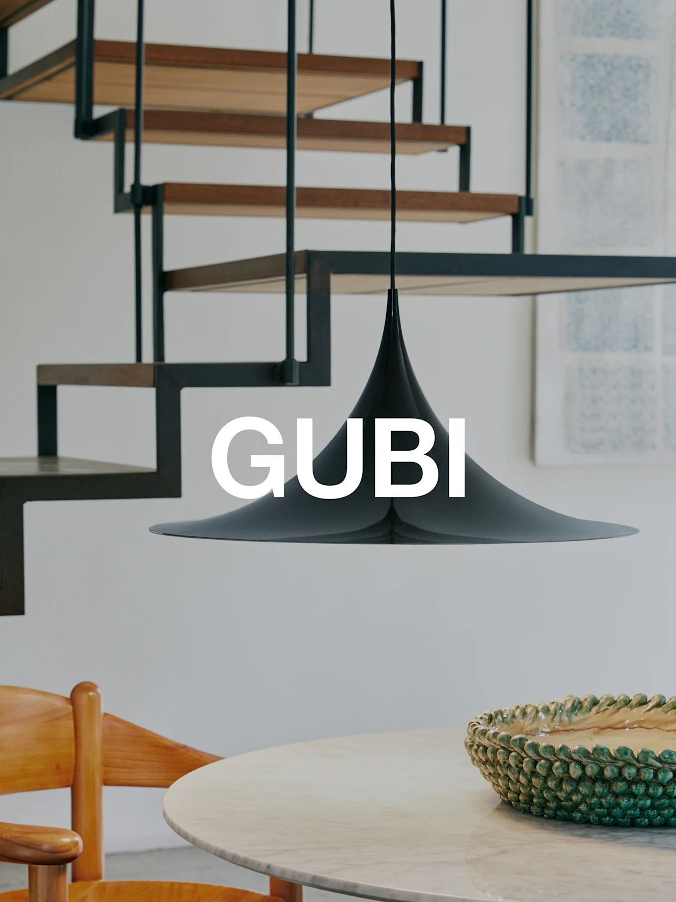 Une lampe suspendue conique noire au-dessus d'une table en marbre avec un bol vert et une chaise en bois. Un escalier moderne en arrière-plan. Texte 'GUBI'.