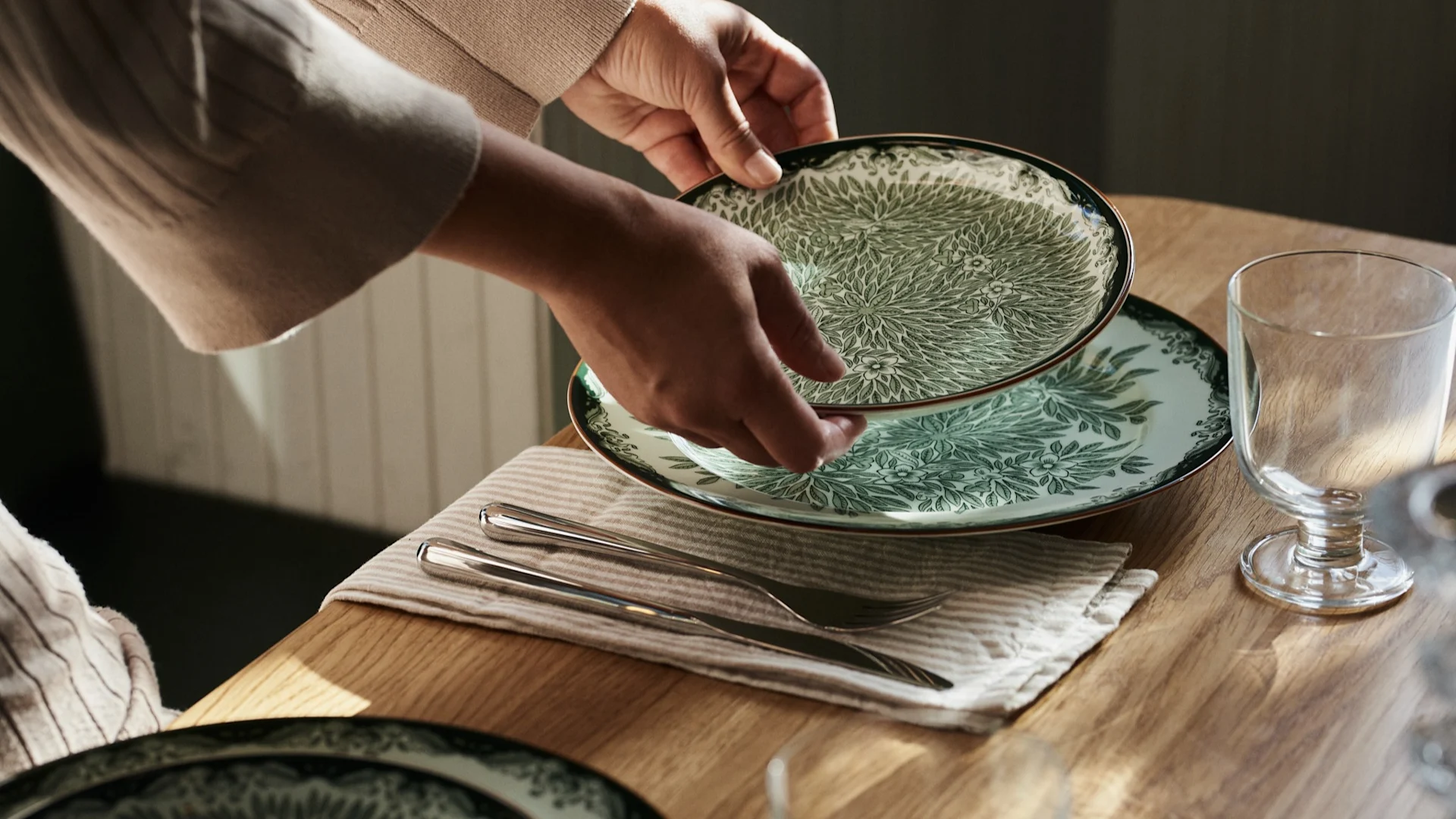 Des mains placent une assiette à motifs verts et blancs sur une table en bois avec des couverts et un verre.