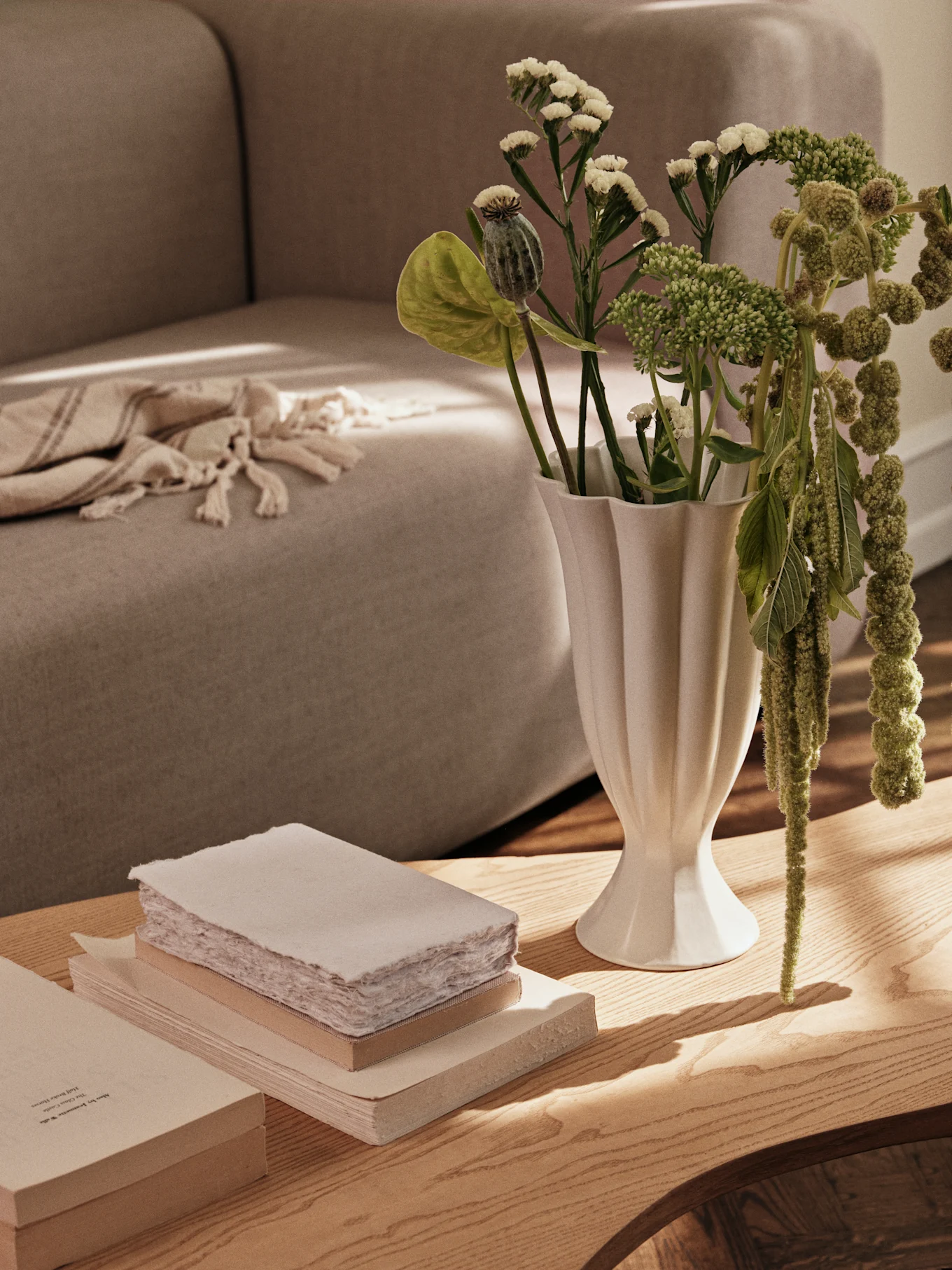 Un vase blanc cannelé avec des plantes vertes et une pile de livres sur une table basse en bois, avec un canapé en arrière-plan.