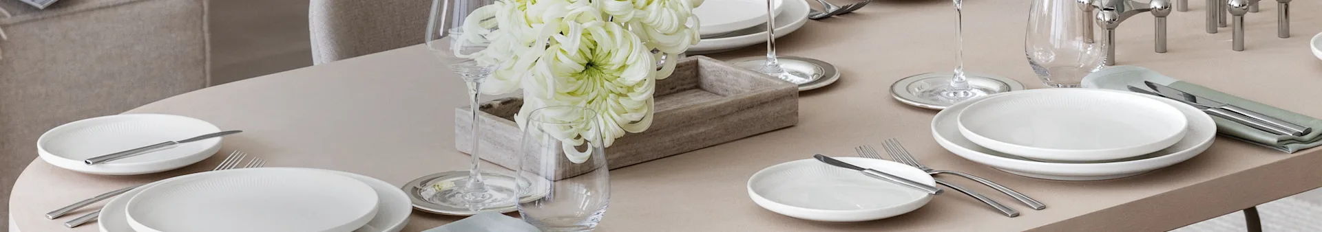 Une table à manger élégamment dressée avec des assiettes blanches, des couverts argentés, des verres à vin et à eau, et un centre de table de grandes fleurs blanches dans une boîte en bois.