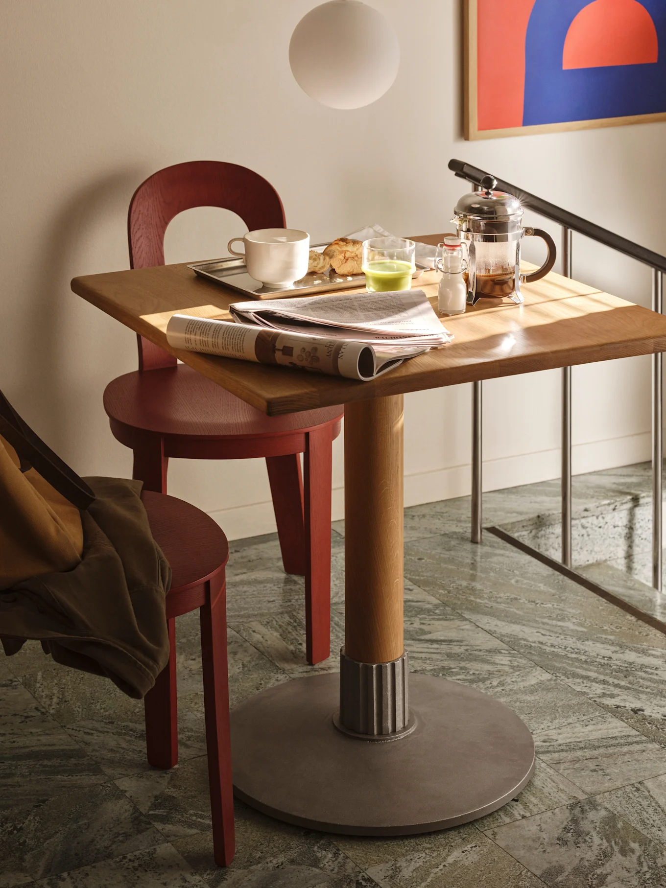 Une table carrée en bois avec petit-déjeuner, journal et cafetière à piston, flanquée de deux chaises rouges sur carrelage.