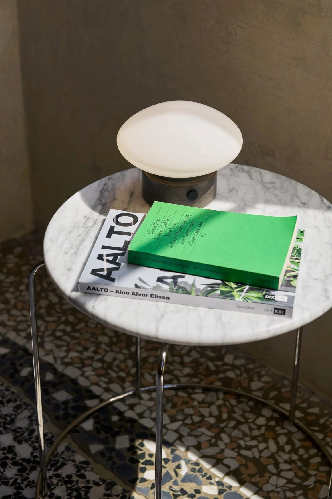 La lampe portable Mist AP12 de &tradition est posée sur une petite table ronde en marbre, à côté de quelques livres. 