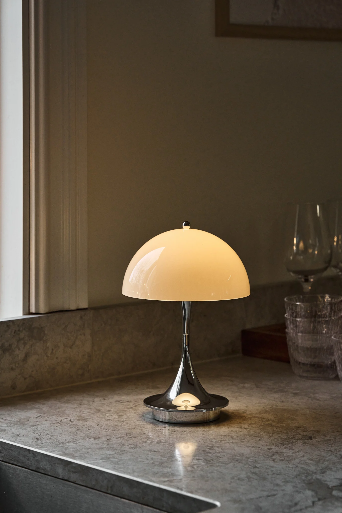 Lampe de table en chrome poli avec un abat-jour dôme crème lumineux sur un comptoir gris moucheté, à côté d'une fenêtre et de verres flous.