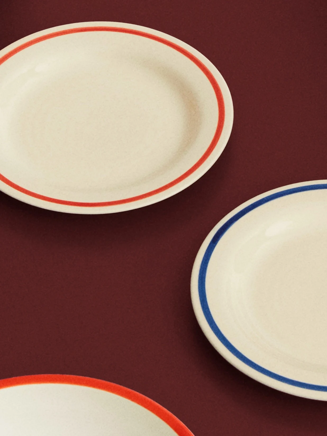 Trois assiettes ovales crème avec des bords rouges et bleus sur une surface rouge foncé.
