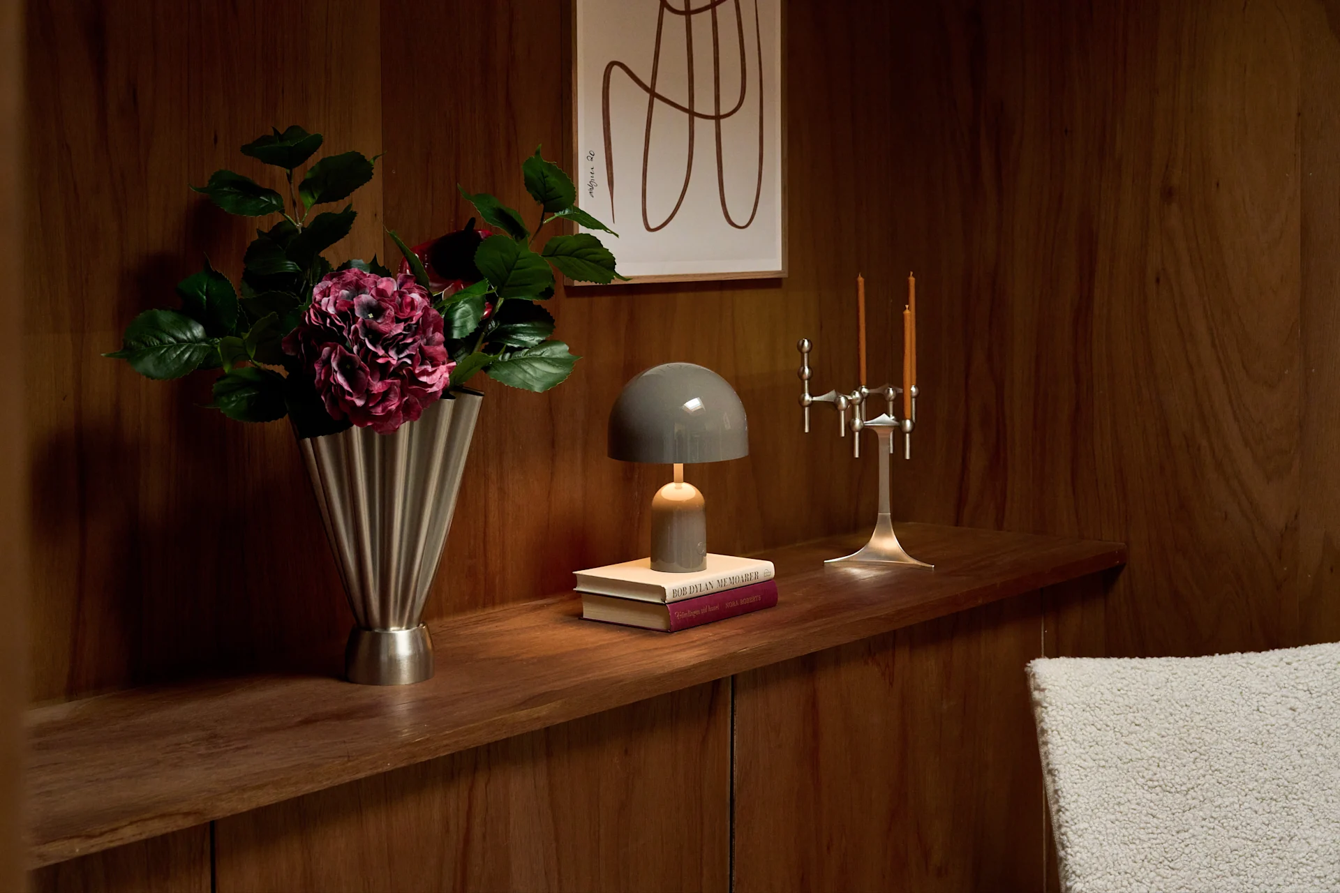 Étagère en bois: vase cannelé argenté et fleurs violettes, lampe champignon grise sur livres, candélabre argenté et deux bougies.