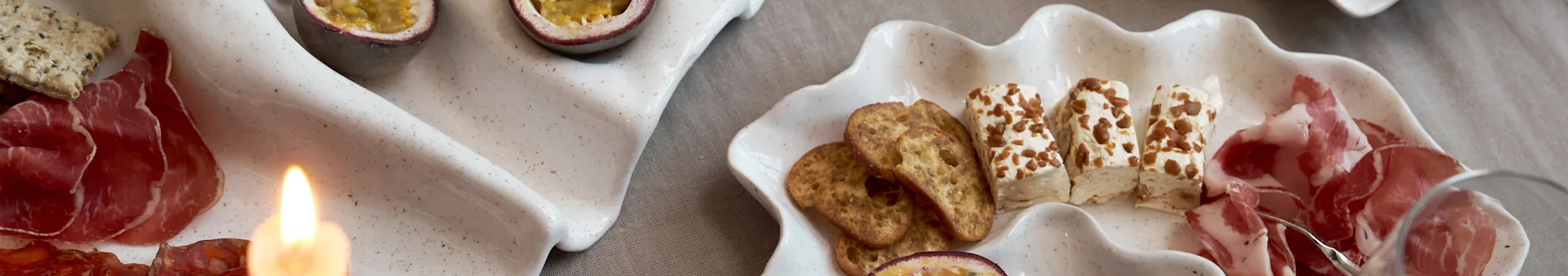Un assortiment d'amuse-gueules sur deux plateaux en forme de coquille avec charcuterie, crackers, fromage et fruits de la passion, avec une bougie allumée.