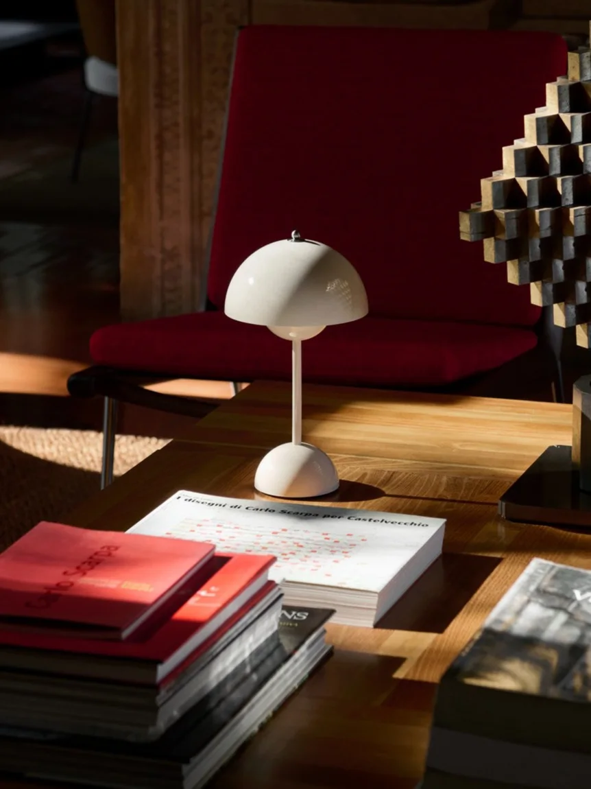 Une lampe de table blanche en forme de champignon, des livres et une sculpture géométrique sur une table basse en bois, avec un fauteuil rouge derrière.