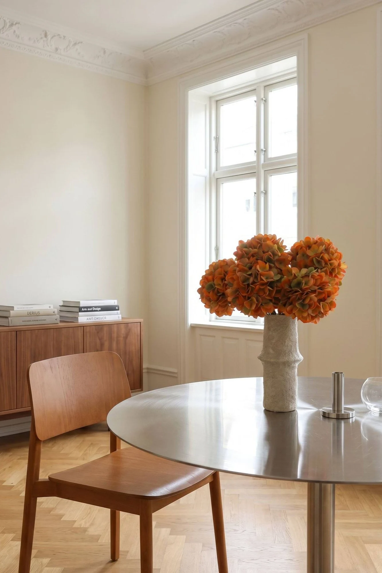 Une salle à manger lumineuse avec une table ronde en métal, une chaise en bois et un vase de fleurs oranges.