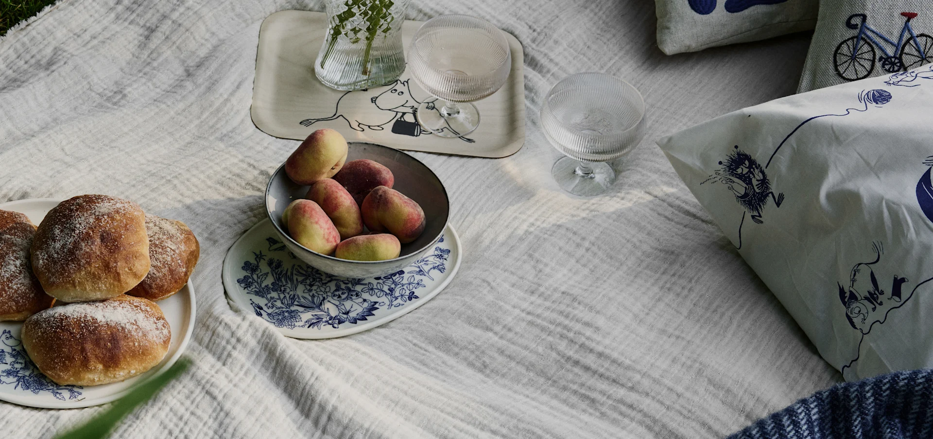 Scène de pique-nique sur une couverture blanche avec des petits pains, des pêches, des boissons et des coussins à motifs Moomin et vélo.