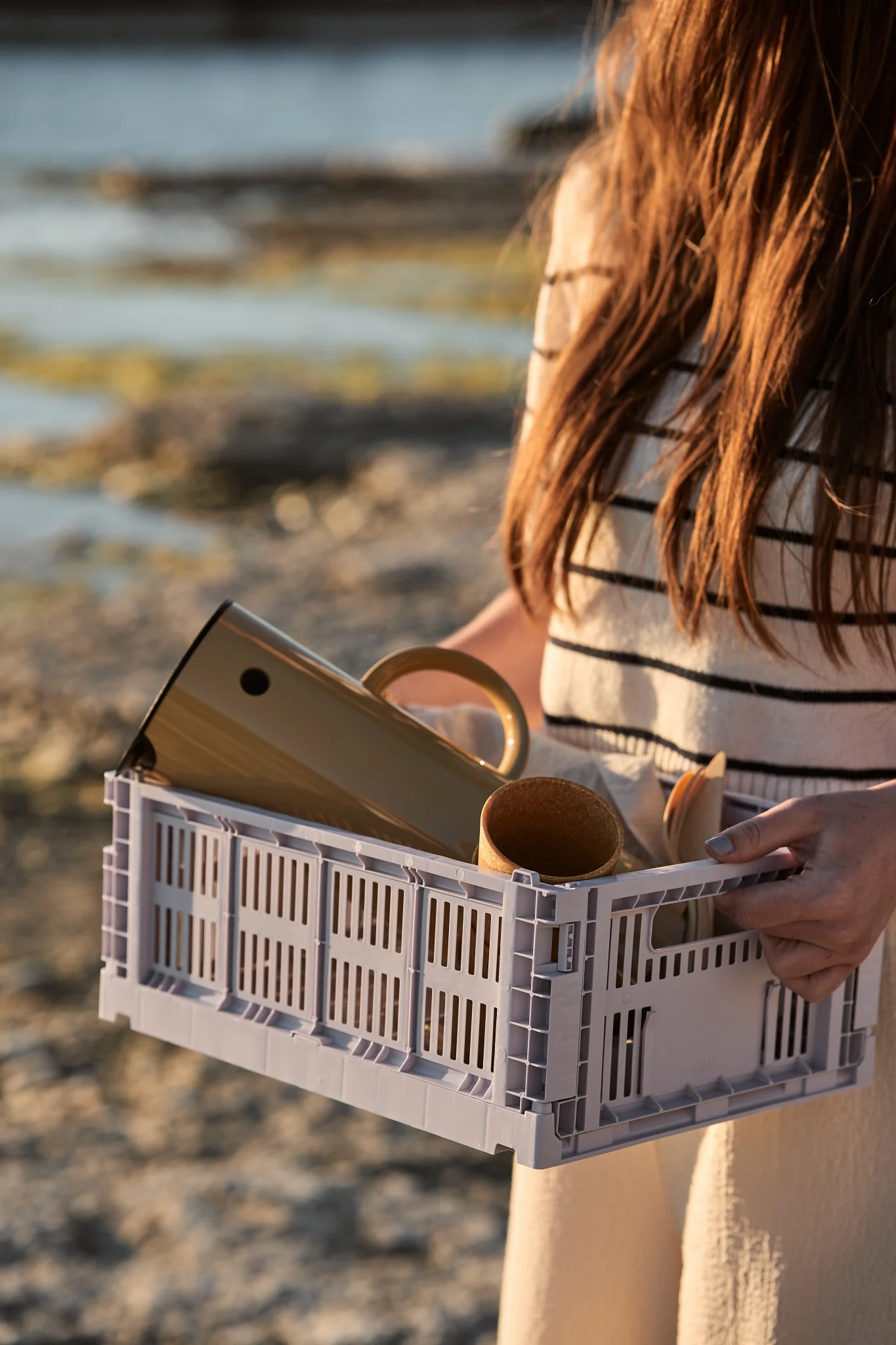 Une personne tient une caisse à lamelles gris clair contenant une bouteille isotherme beige, un gobelet et des couverts en bois, avec la plage en arrière-plan.