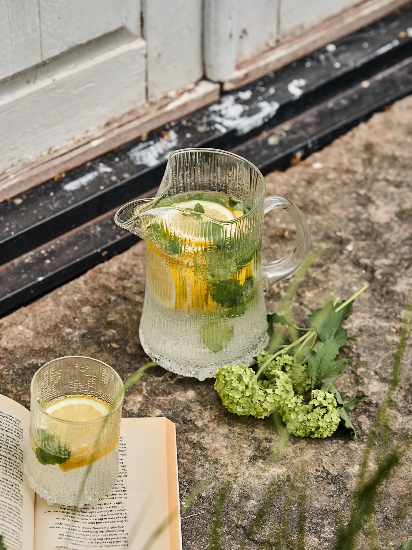 Pichet en verre nervuré et verre assorti avec de l'eau citronnée et mentholée, à côté d'un livre ouvert et de fleurs vertes sur la pierre.