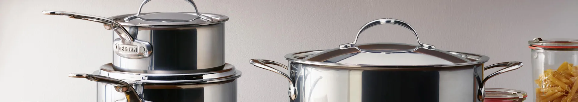 Casseroles en acier inoxydable avec couvercles, une grande à droite et d'autres empilées à gauche, à côté d'un pot de pâtes.