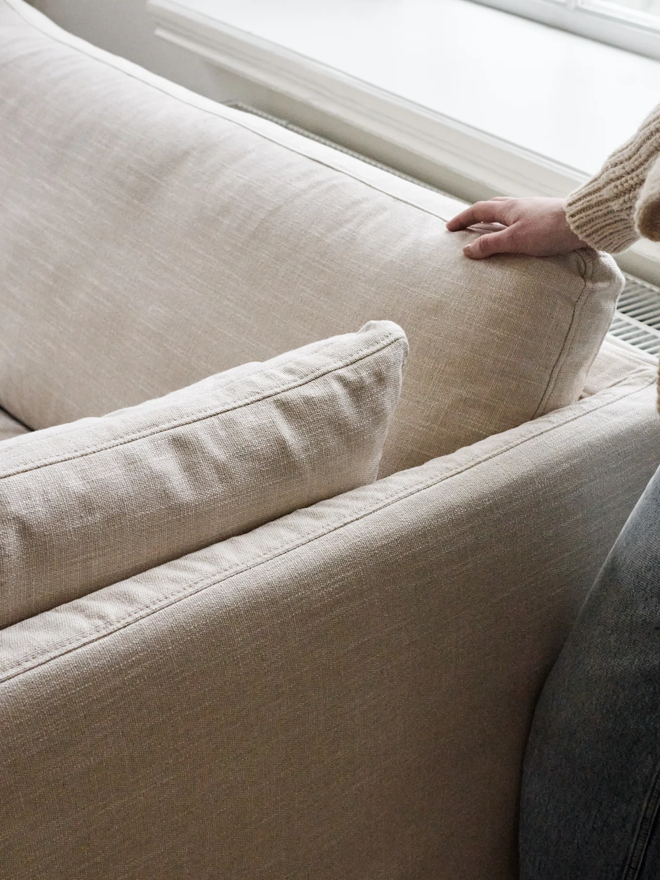 Une main vêtue d'un pull en grosse maille presse doucement le coussin du dossier d'un canapé beige clair.