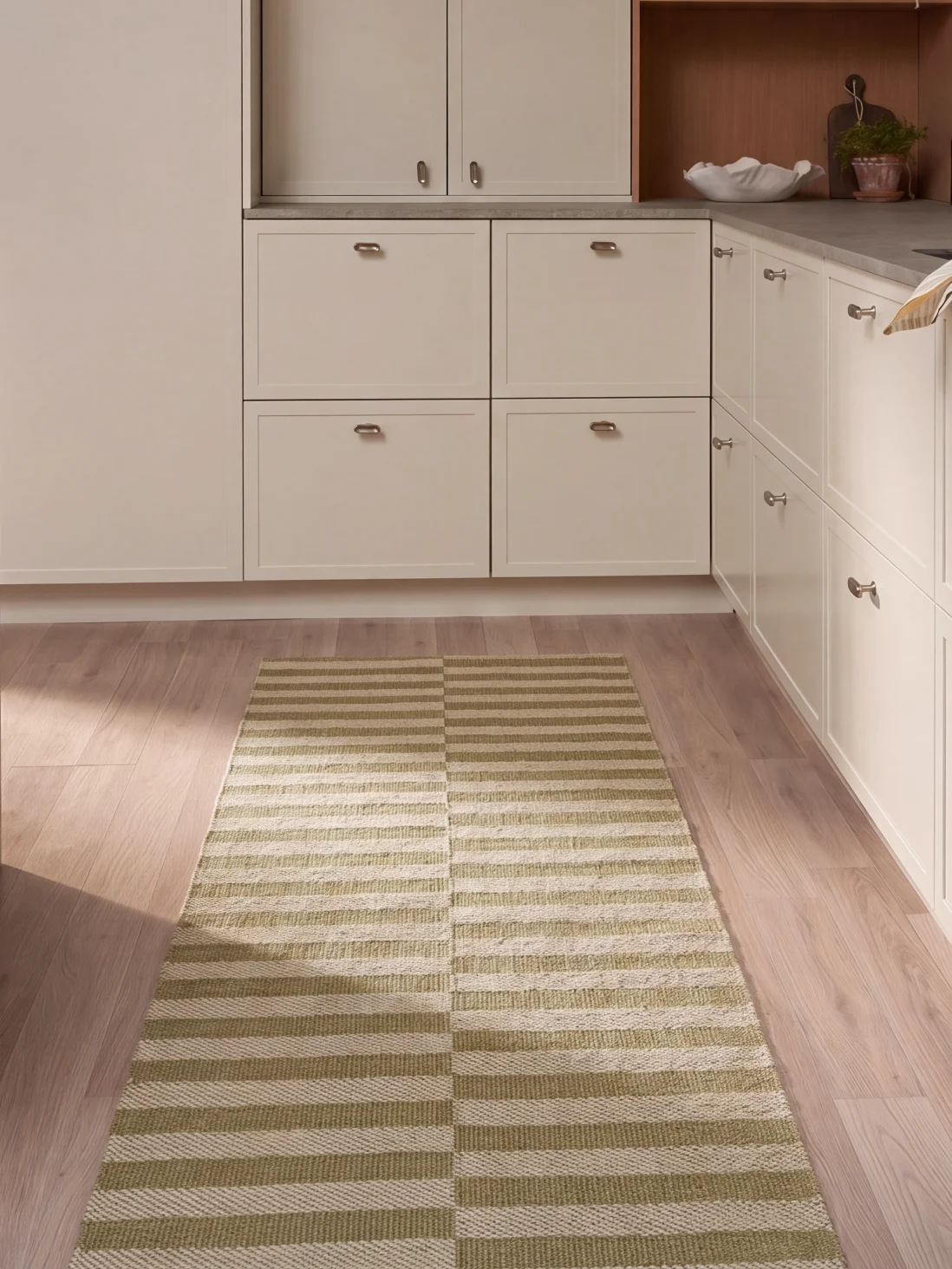 Une cuisine avec des armoires claires, un sol en bois et un long tapis rectangulaire à rayures horizontales vertes et blanches.