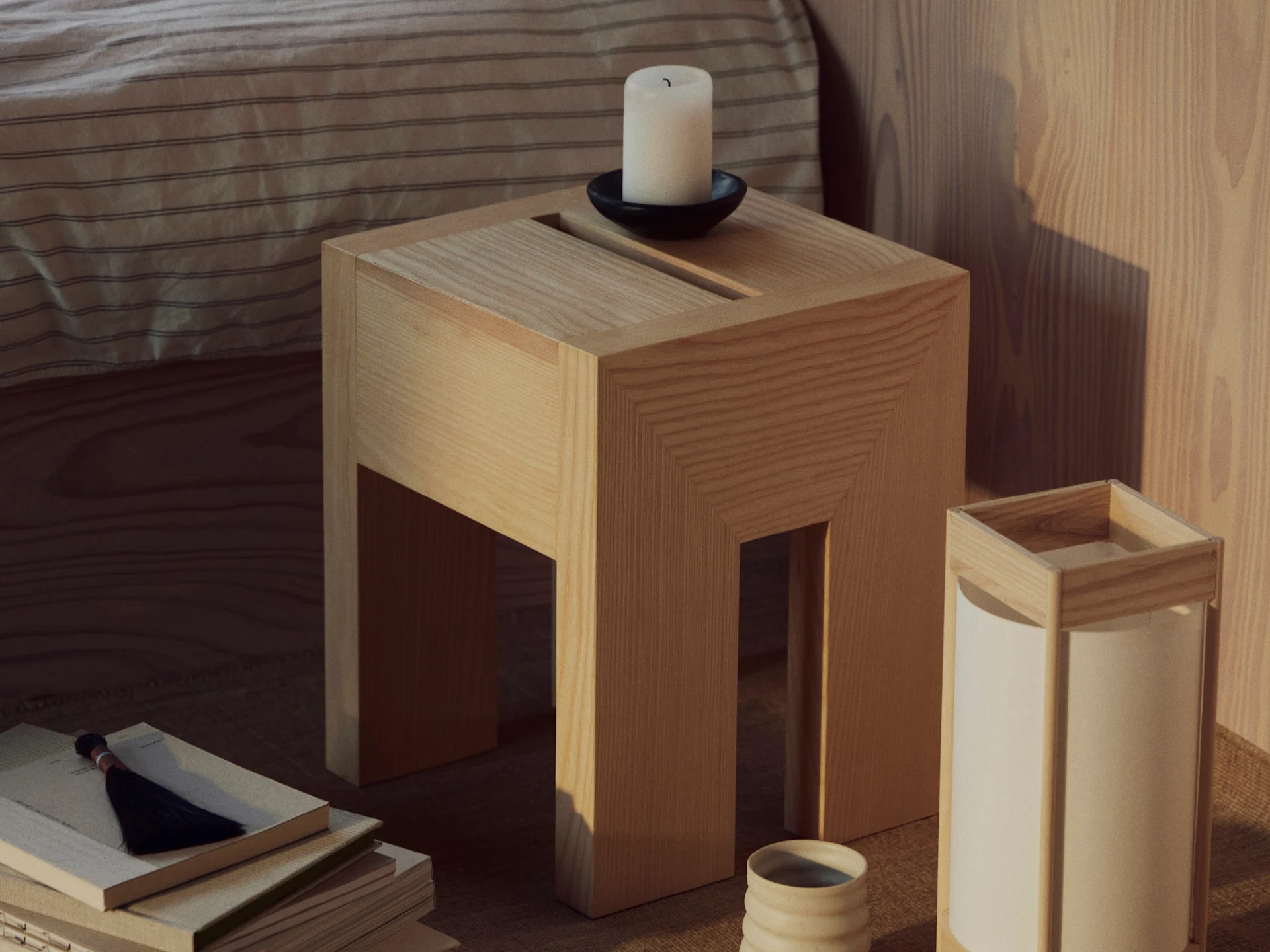 Une table d'appoint en bois avec une bougie blanche dans un support noir, une pile de livres à pompon et une lampe avec tasse en céramique.