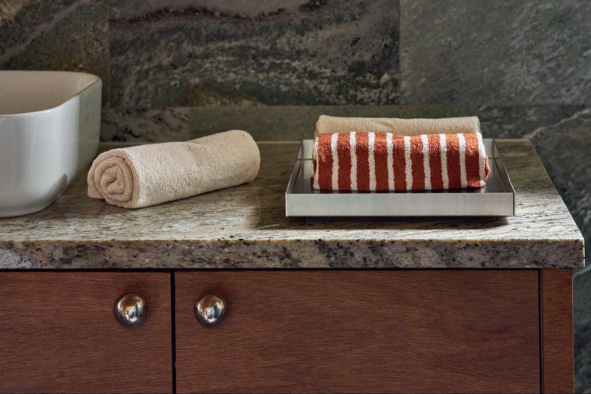 Serviettes roulées beiges et rayées couleur rouille sur un plateau argenté sur un comptoir en granit, à côté d'un lavabo blanc et de meubles en bois.