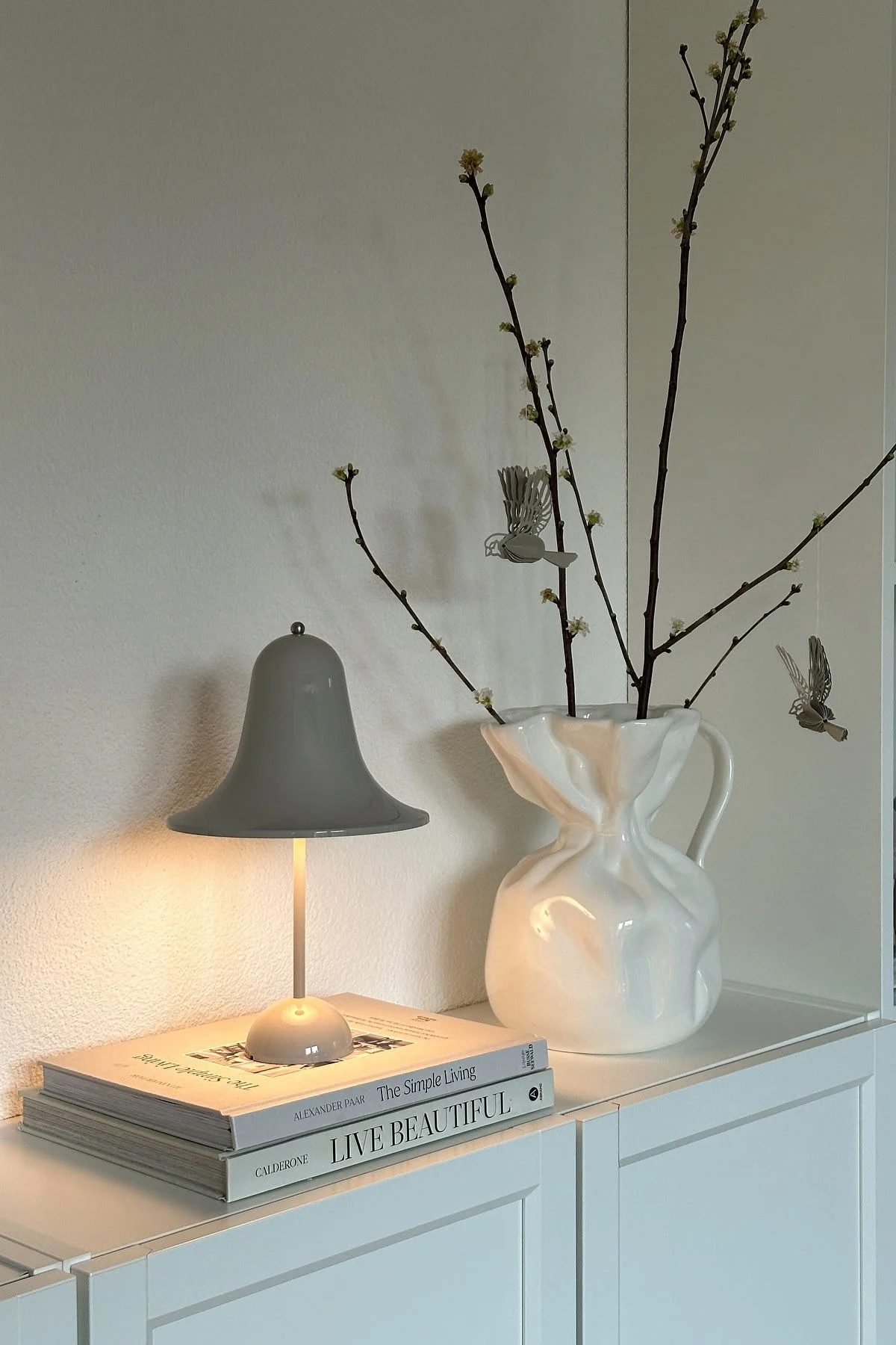 Sur @oursmall.home, vous pouvez voir deux cadeaux possibles pour la fête des mères : la lampe de table Pantorp de Verpan est posée sur une commode, à côté de laquelle se trouve le vase Crumple de Byon avec quelques brindilles.