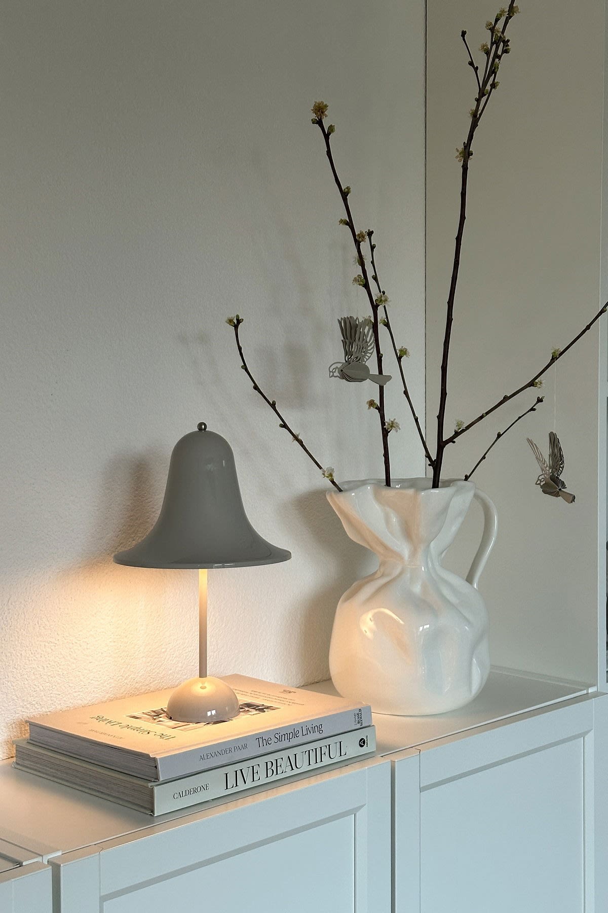Sur @oursmall.home, vous pouvez voir deux cadeaux possibles pour la fête des mères : la lampe de table Pantorp de Verpan est posée sur une commode, à côté de laquelle se trouve le vase Crumple de Byon avec quelques brindilles.