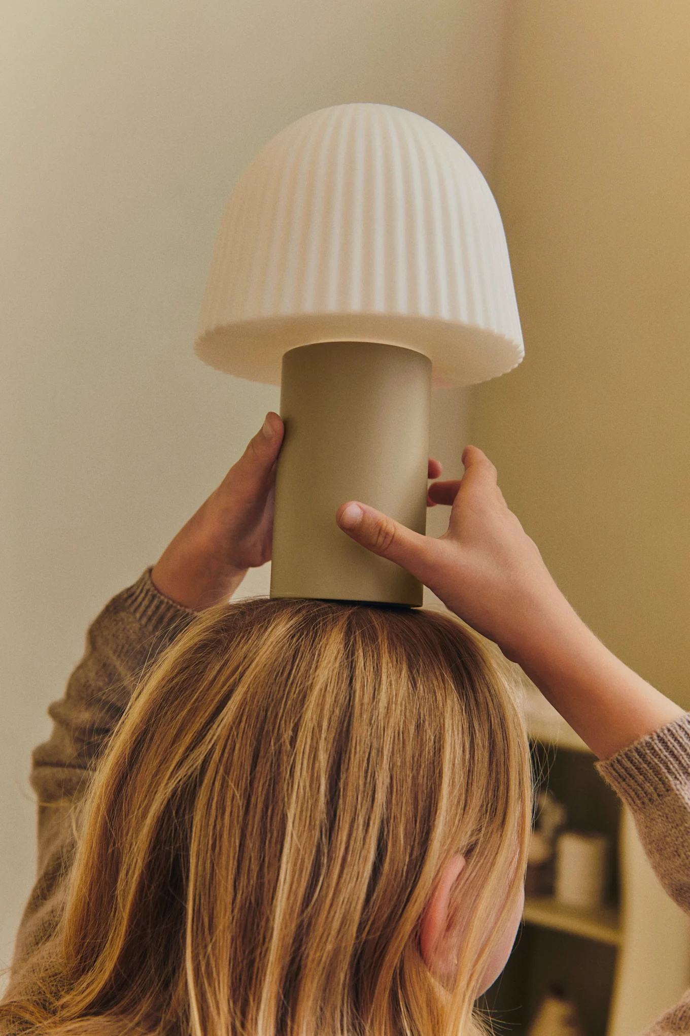 Enfant de dos, équilibrant une lampe champignon verte à abat-jour blanc nervuré sur sa tête.