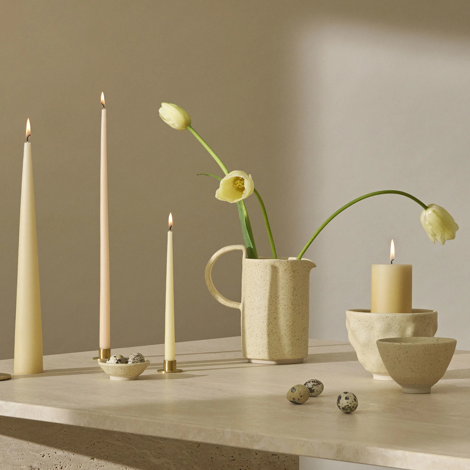 Une nature morte avec des bougies allumées, des tulipes jaune pâle dans un pichet moucheté et des œufs de caille sur une surface texturée.