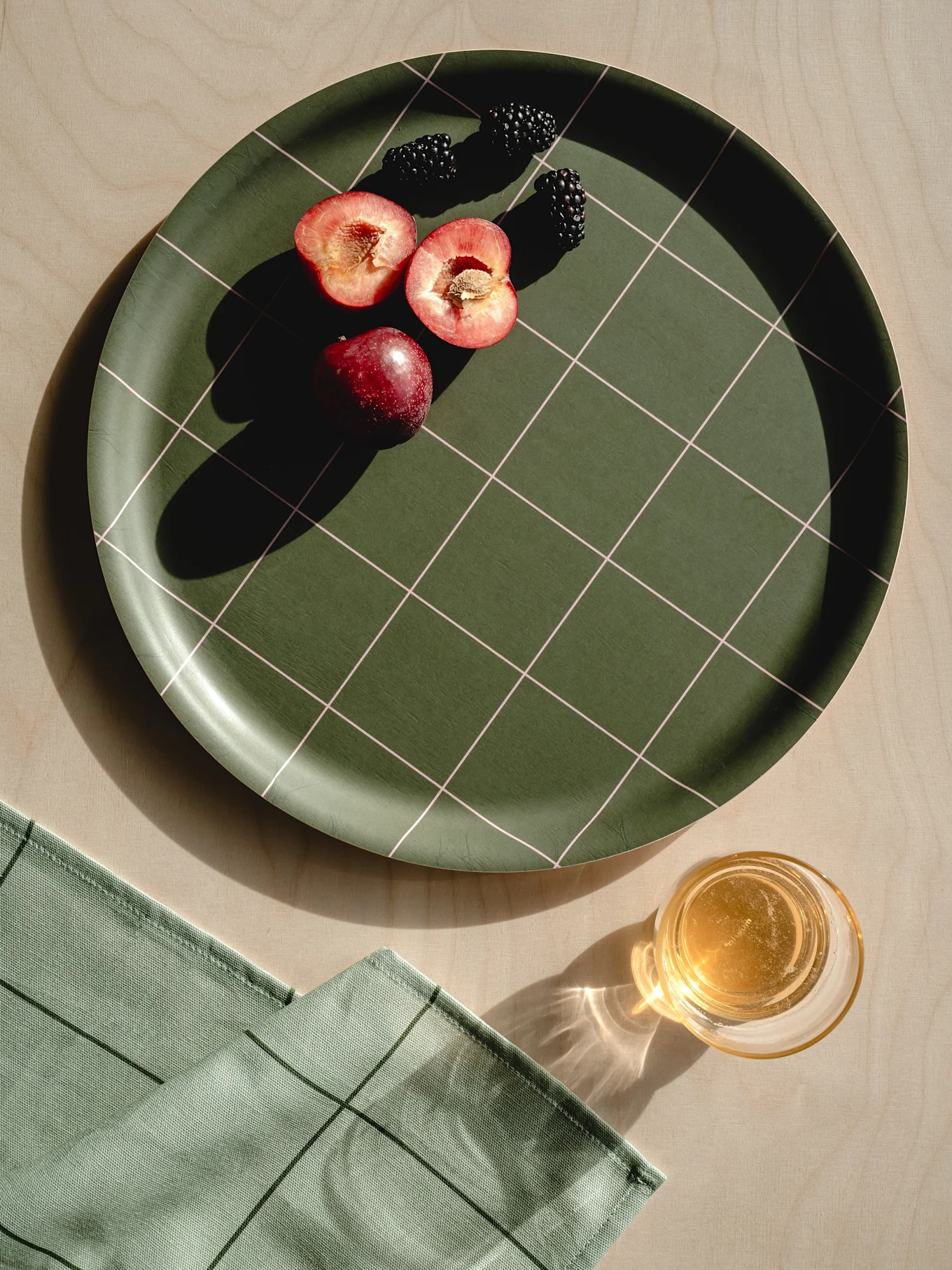 Plateau à carreaux vert foncé avec prunes rouges, mûres, serviette vert clair et verre de jus sur bois.