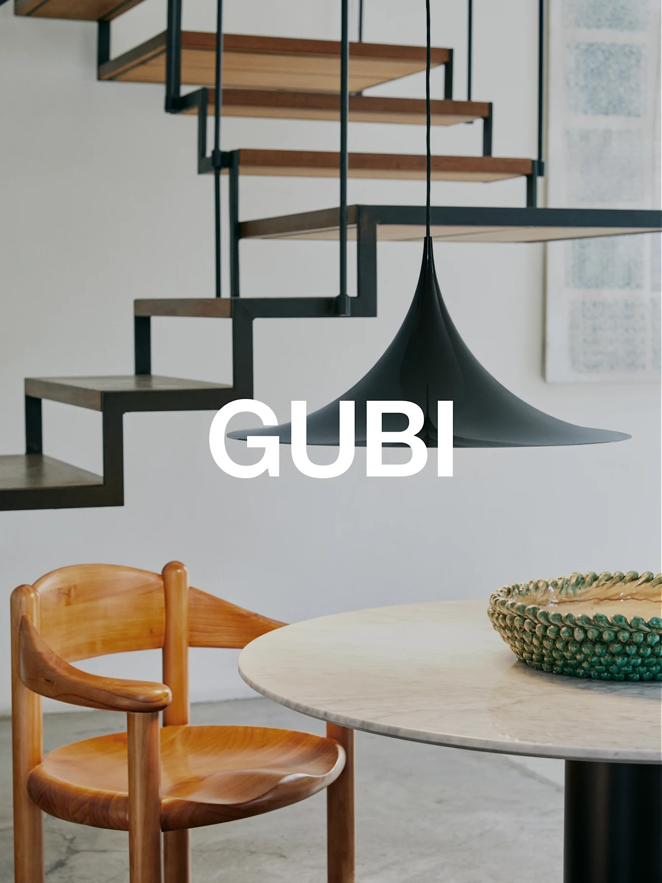 Logo GUBI dans un intérieur moderne avec chaise en bois, table en marbre, bol vert, suspension noire et escalier moderne.