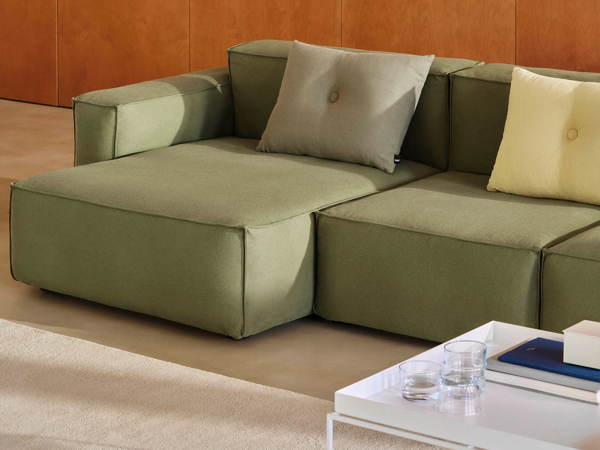 Un canapé modulaire vert olive avec des coussins décoratifs gris clair et jaunes, à côté d'une table basse blanche avec des livres.