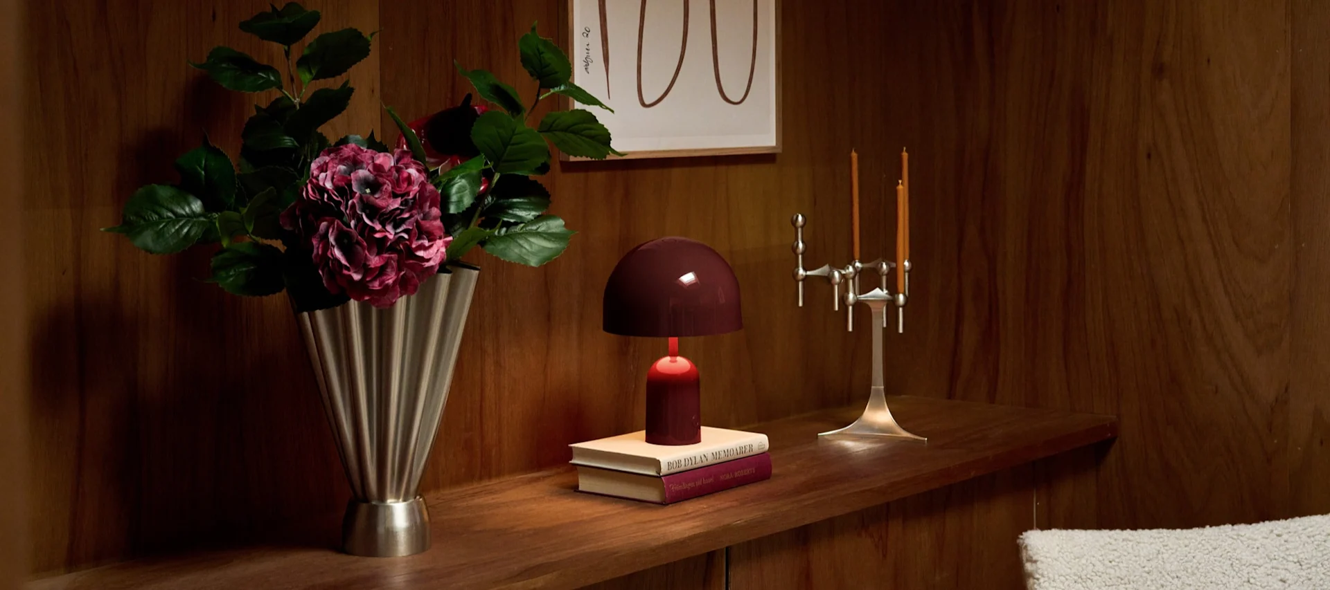 Étagère bois: vase argenté, hortensias foncés, lampe dôme bordeaux sur livres, chandelier argenté avec bougies oranges.