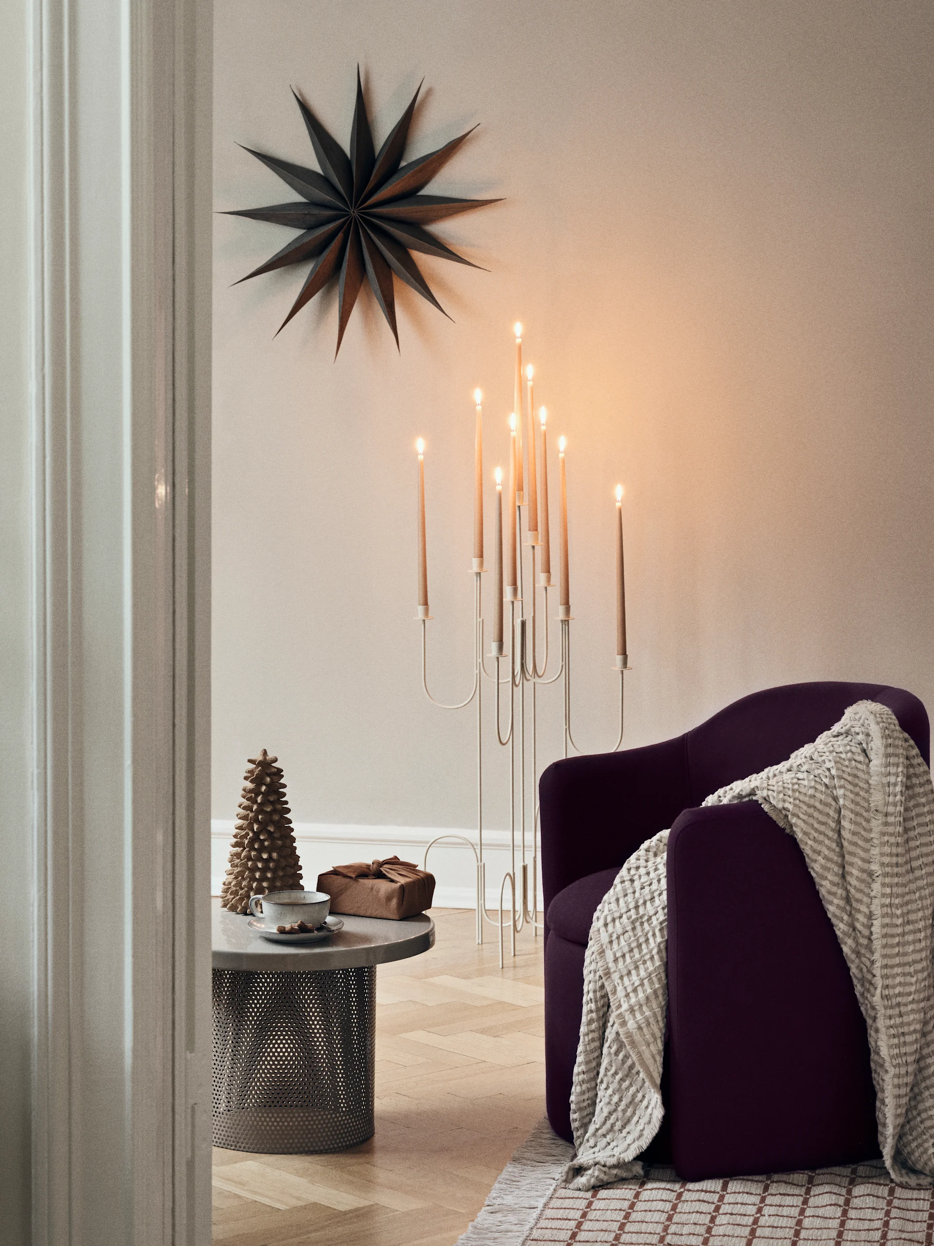 Une scène d'hiver lumineuse à travers une fenêtre, avec des arbres enneigés et un rayon de soleil. À l'intérieur, une table avec des bougies et des sapins en papier, et une étoile en papier est suspendue à la fenêtre.