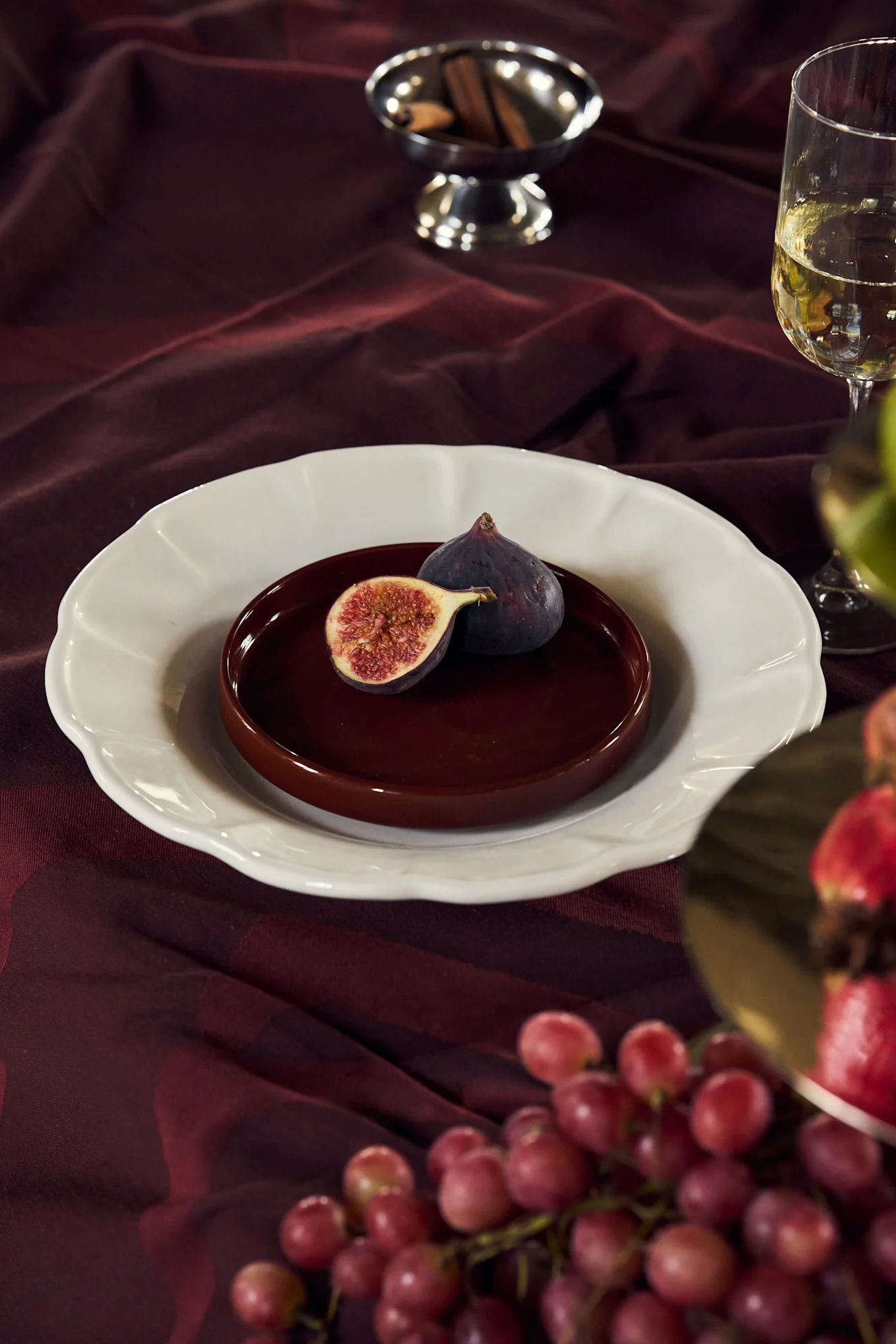 Figues sur une assiette posée sur une nappe bordeaux, avec du vin, des raisins et des bâtons de cannelle.