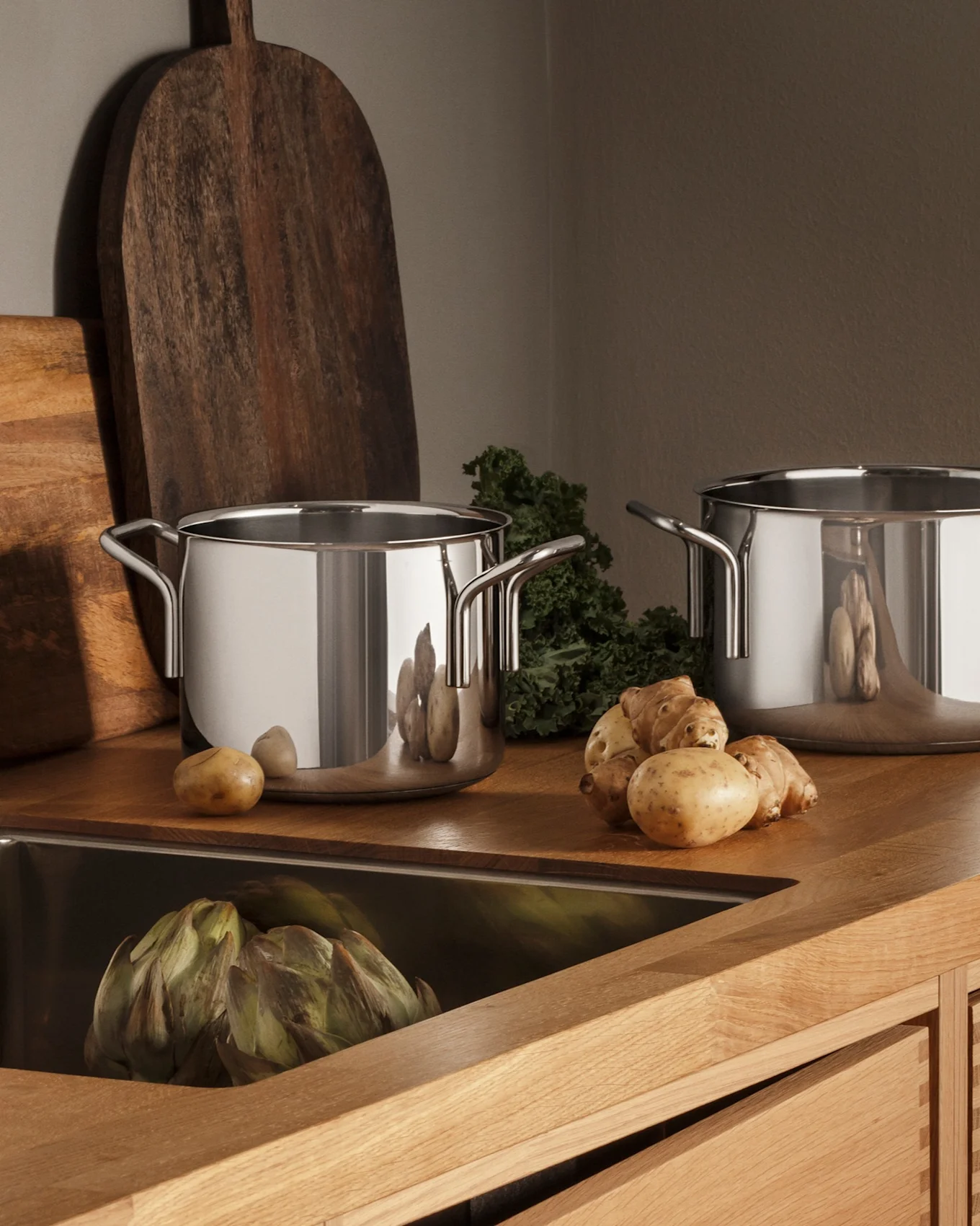 Cuisine moderne avec comptoir en bois, évier en acier inoxydable, robinet, trois casseroles et produits frais comme des pommes de terre et des artichauts.