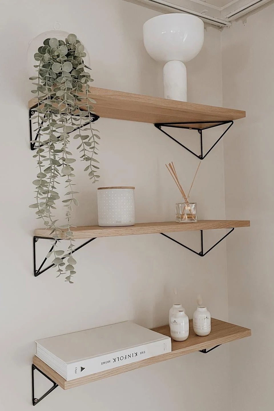 Trois étagères suspendues en bois clair, dotées de supports triangulaires noirs. Parmi les objets, on trouve une plante suspendue, un vase blanc, un récipient à pois, un diffuseur, un livre blanc et de petits vases blancs.