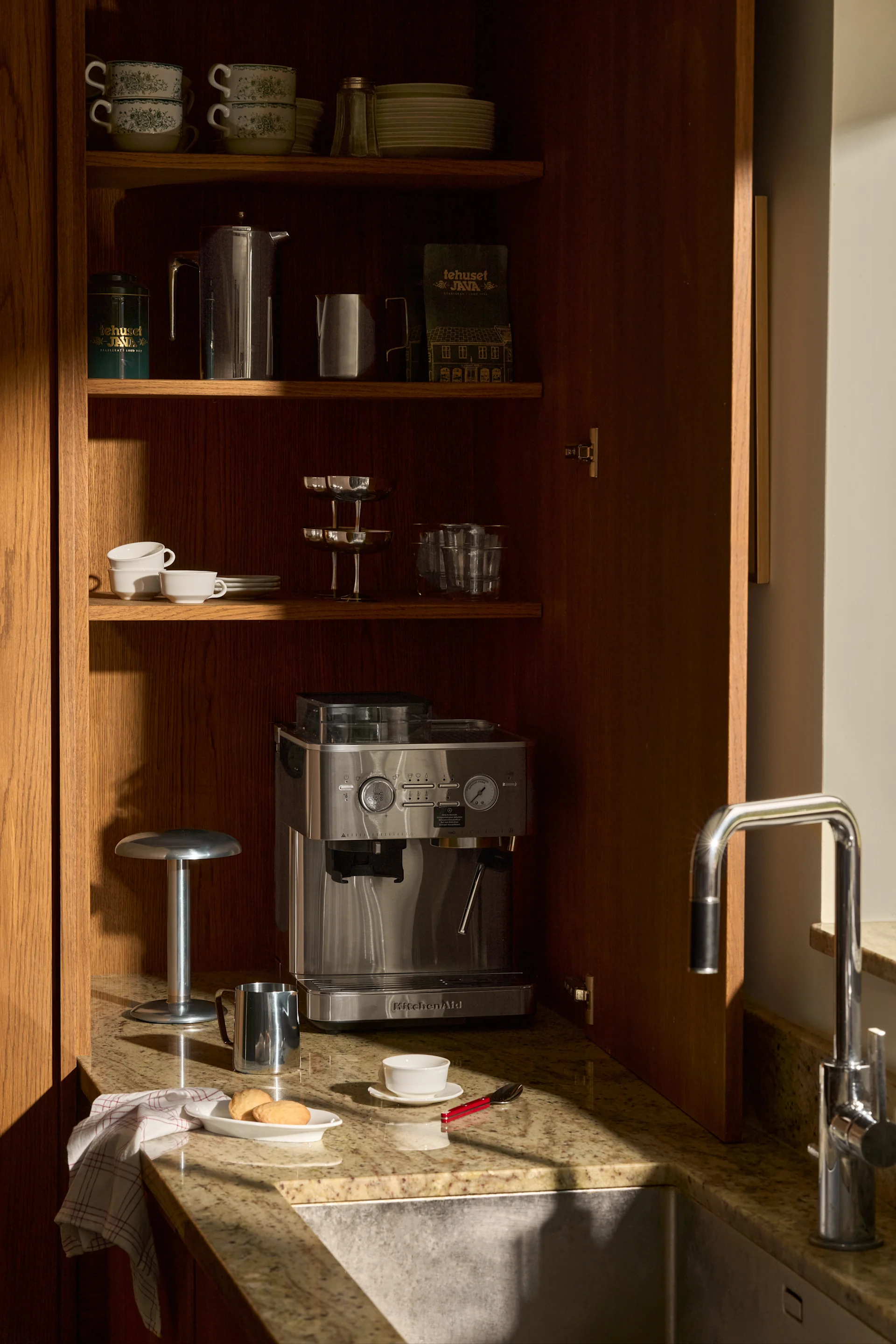 Armoire en bois ouverte: machine à expresso KitchenAid, tasses, assiettes, café et évier sur comptoir de granit.