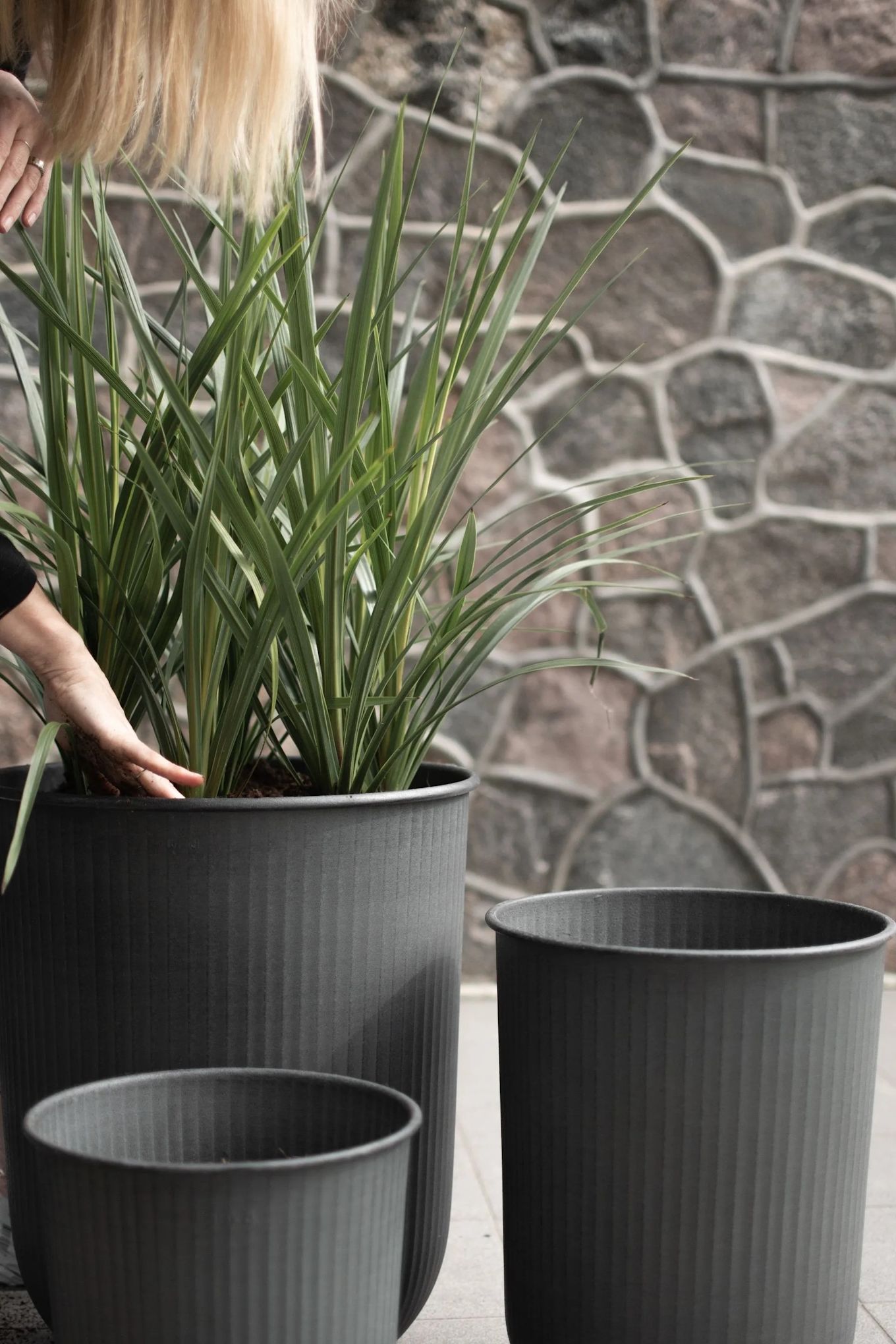 Une femme se penche pour planter une herbe haute dans un grand pot métallique pour l'extérieur - Out from DBKD