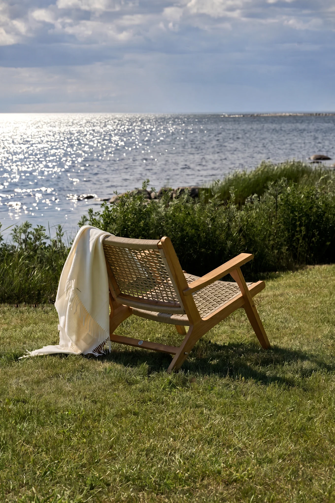 Fauteuil de jardin en bois avec un plaid blanc sur l'herbe verte, face à l'eau scintillante sous un ciel nuageux.