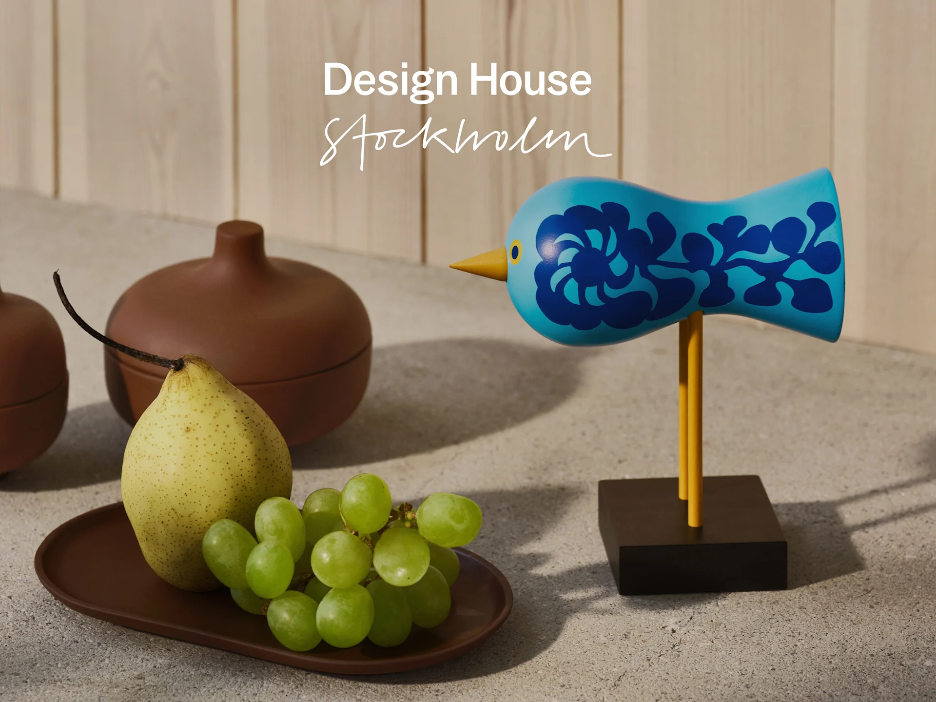 Nature morte: figurine d'oiseau bleu, poire, raisins verts et pot marron sur béton, avec 'Design House Stockholm'.