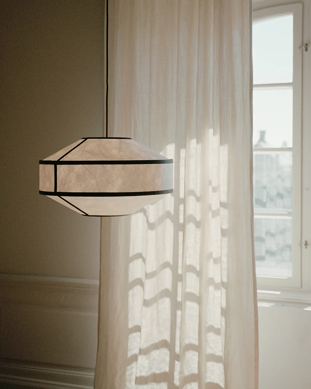 Une lampe suspendue géométrique en papier avec des cadres foncés est suspendue près d'un rideau transparent et d'une fenêtre ensoleillés.